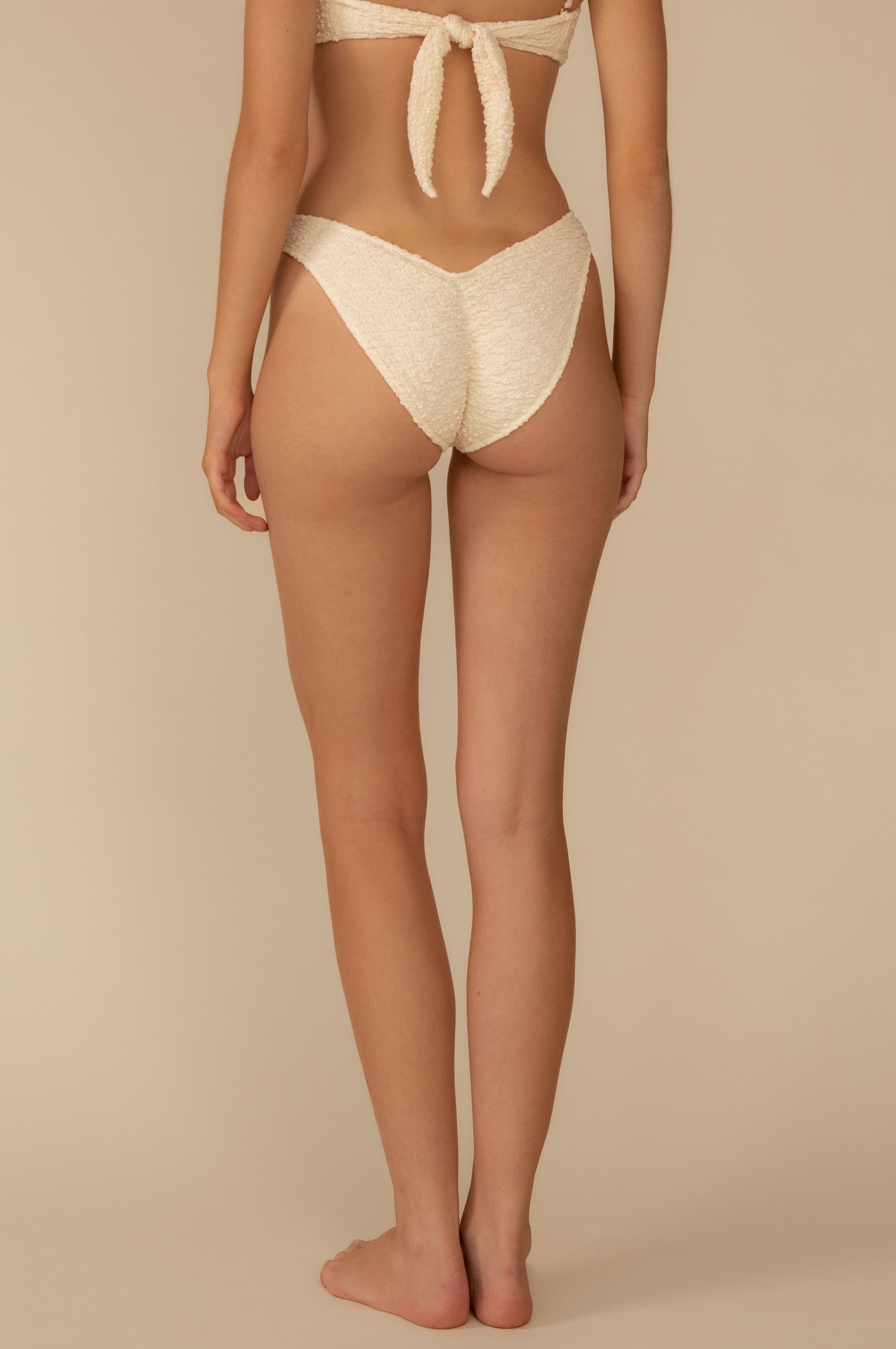 A woman stands barefoot on a neutral backdrop, shown from behind, wearing Montce’s Amalia Texture Lulu Bikini Bottom in cream with a high-waisted fit and paired with a tied back bikini top.