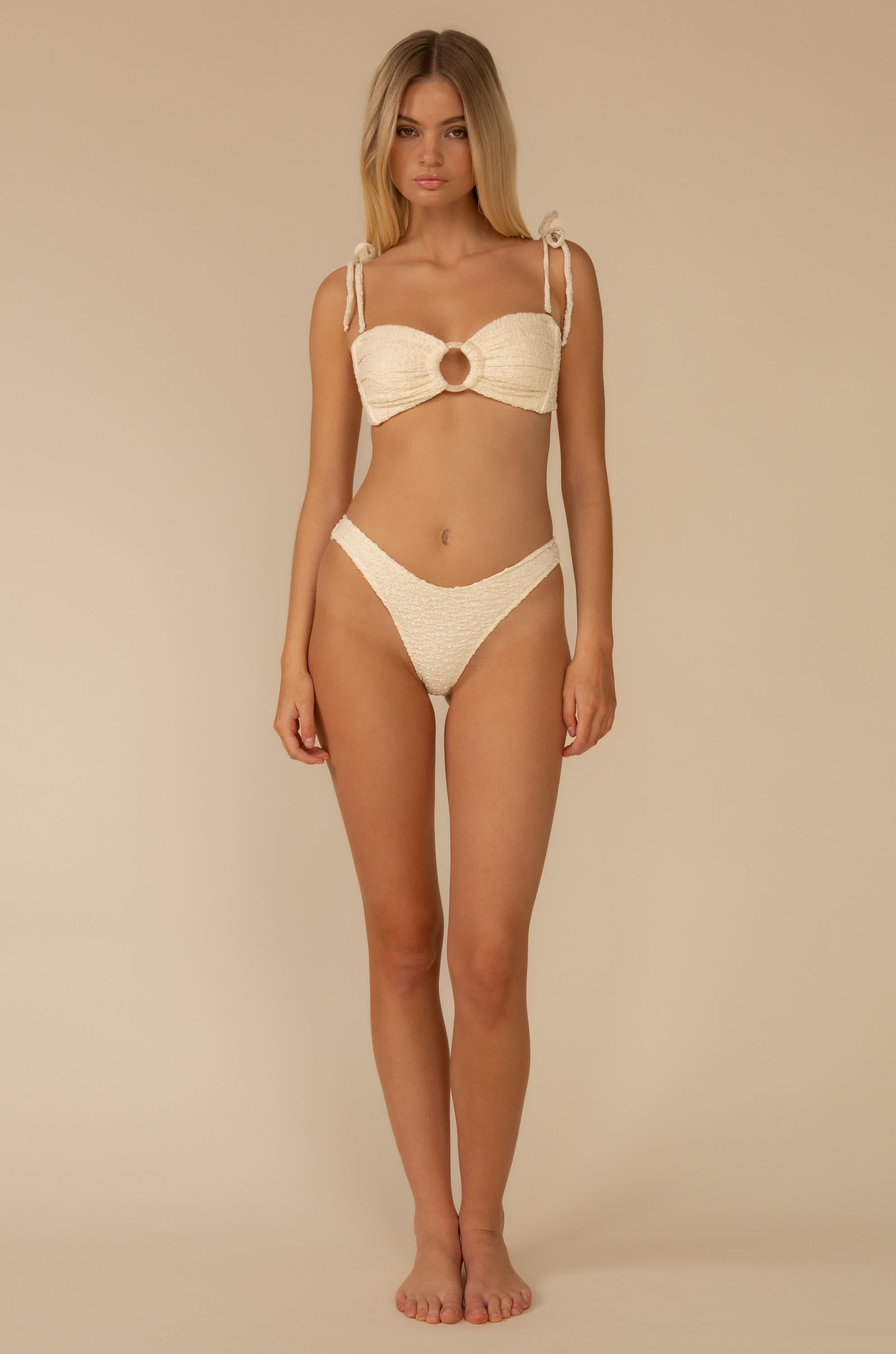 A woman stands barefoot against a beige background wearing the Montce Amalia Texture Lulu Bikini Bottom, featuring a cream-colored textured fabric, circular bust cutout, and tie shoulder straps. She looks directly at the camera with a neutral expression.
