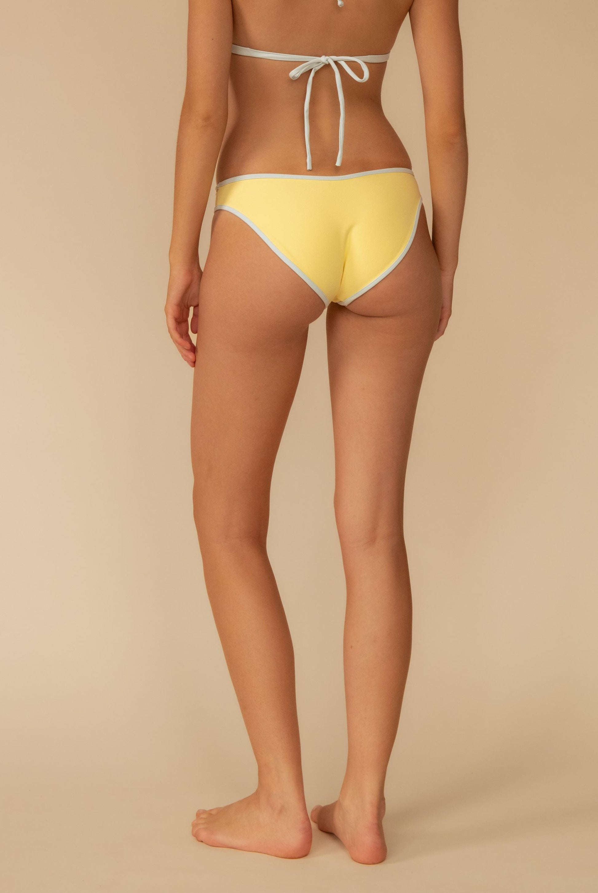 A person is seen from behind wearing the Montce Sunshine Binded Lulu Bikini Bottom, a moderate coverage swimwear in yellow with light blue trim, featuring a bikini top tied at the back with a bow.