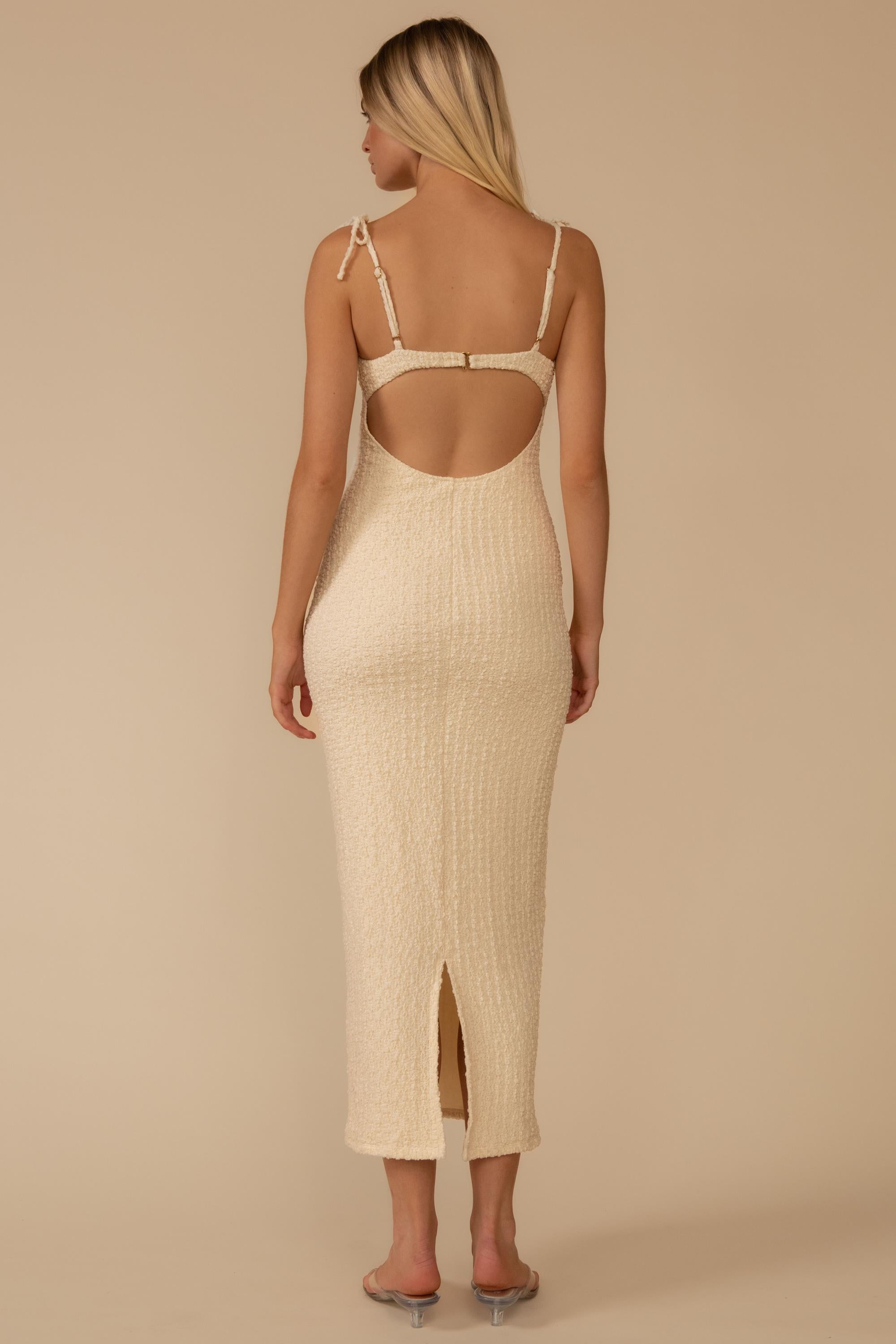 Back view of a woman wearing the Montce Petal Long Slip Dress, a cream textured midi with a low back, high slit, and clear heels.