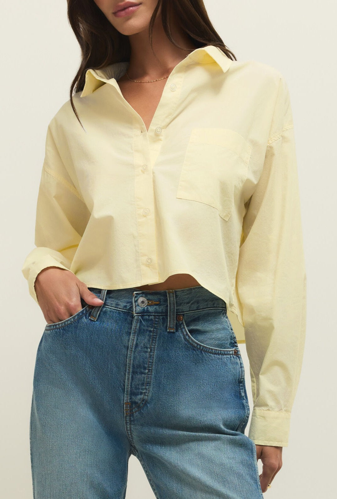 A woman wears the Lisette Top by Z Supply in PINEAPPLE—an oversized, pastel yellow shirt featuring long sleeves, a button-up front, and a chest pocket—paired with high-waisted blue jeans. Her head is partially out of frame.