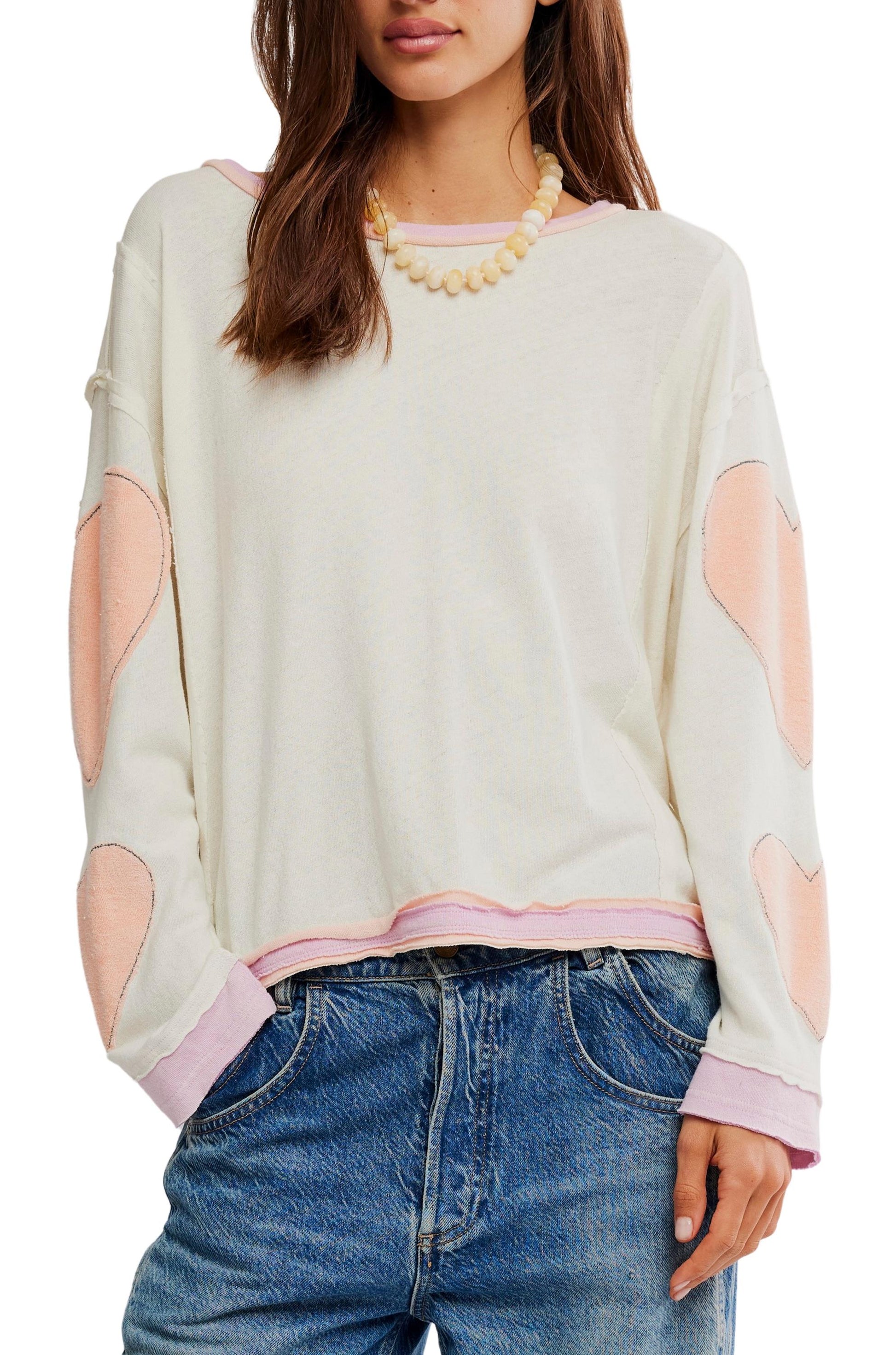 A woman wears the Youre A Star Tee by Free People—a white, oversized long-sleeve top with light pink heart patches and trim—paired with blue jeans and a beaded necklace for an effortless look. Her brown hair is down.