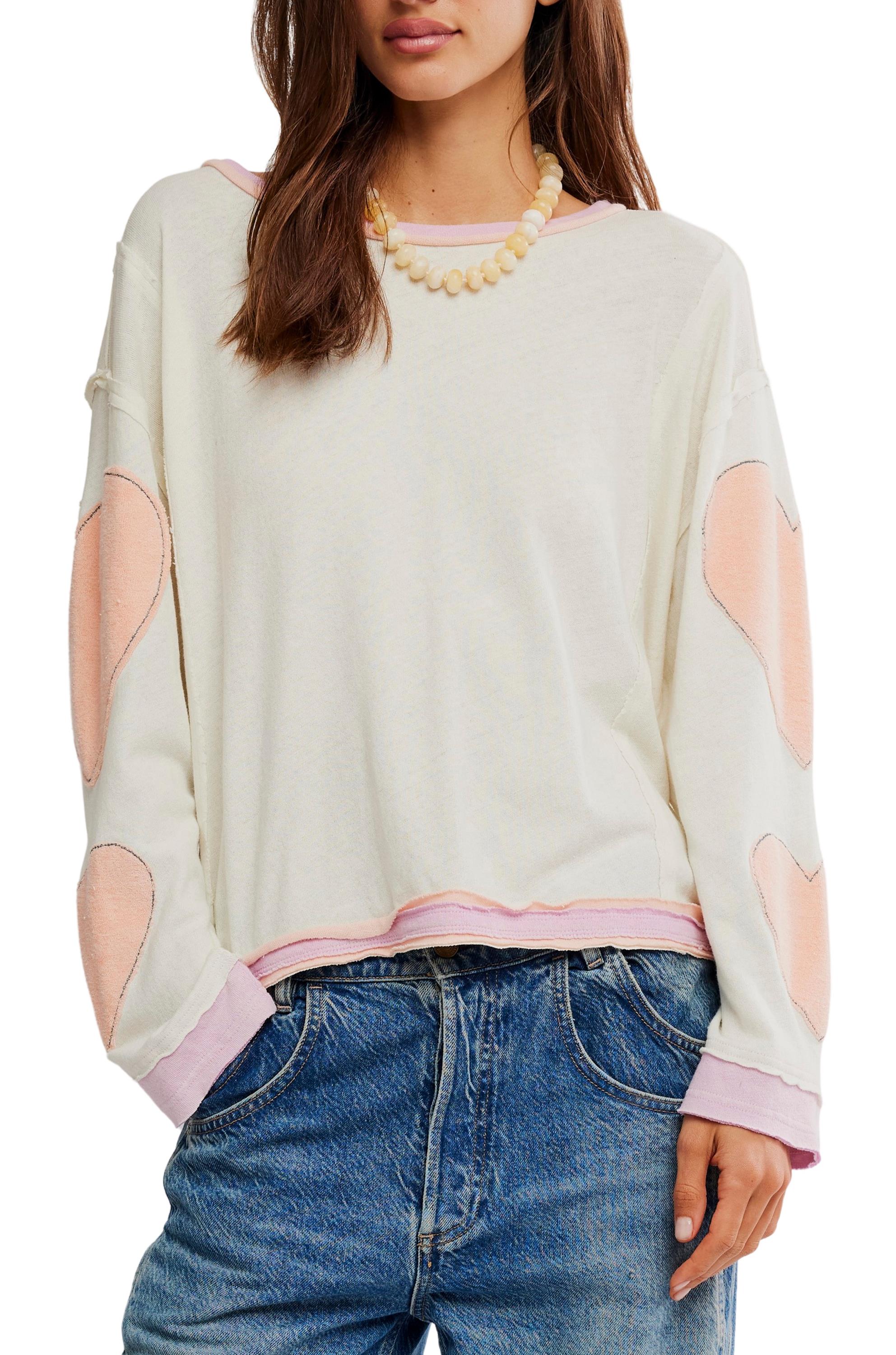 A woman wears the Youre A Star Tee by Free People—a white, oversized long-sleeve top with light pink heart patches and trim—paired with blue jeans and a beaded necklace for an effortless look. Her brown hair is down.