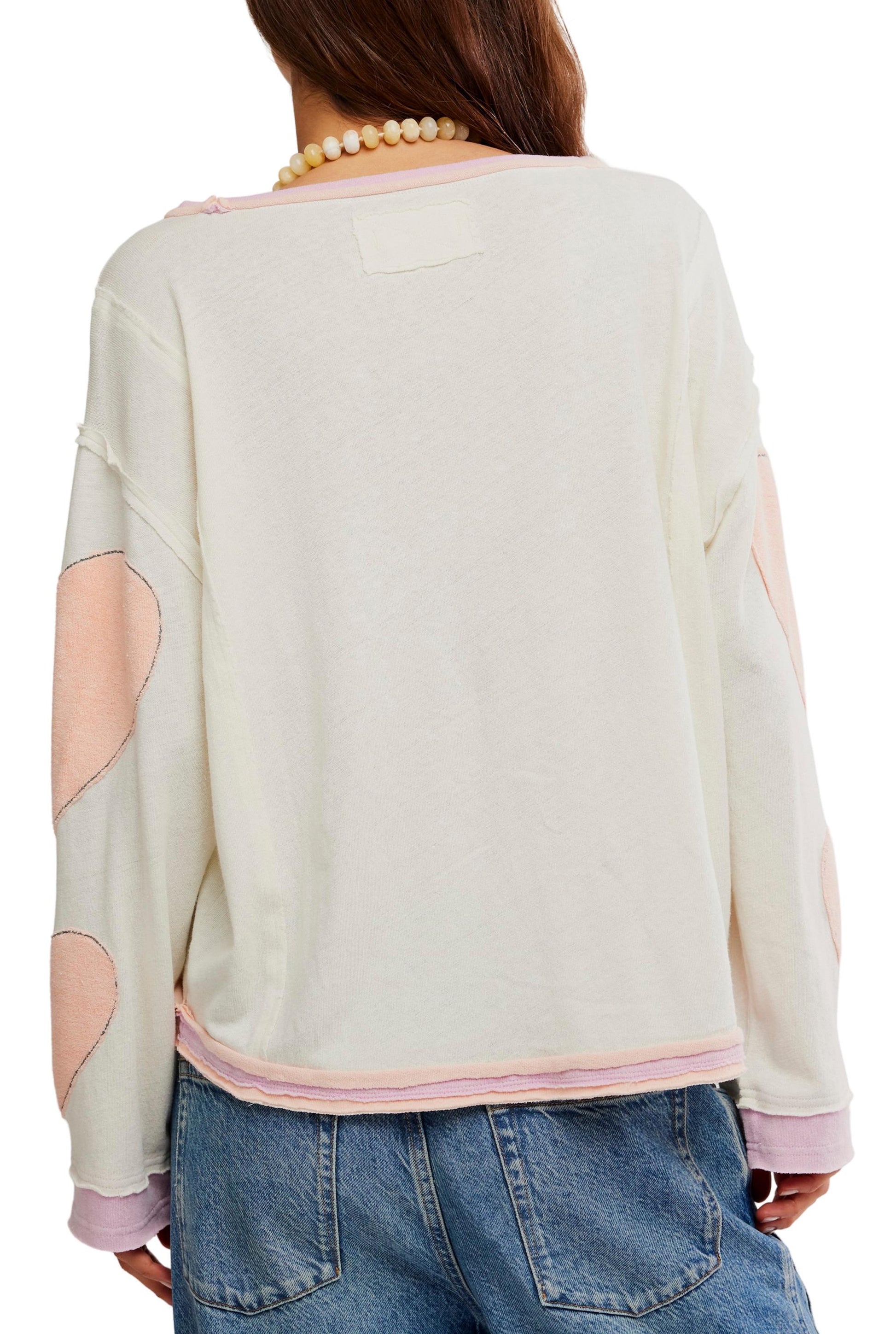 A person with long brown hair, facing away, wears a Free People Youre A Star Tee—an oversized off-white long-sleeve top featuring pink heart patches and subtle star details on the sleeves—paired with blue jeans and a beaded necklace.