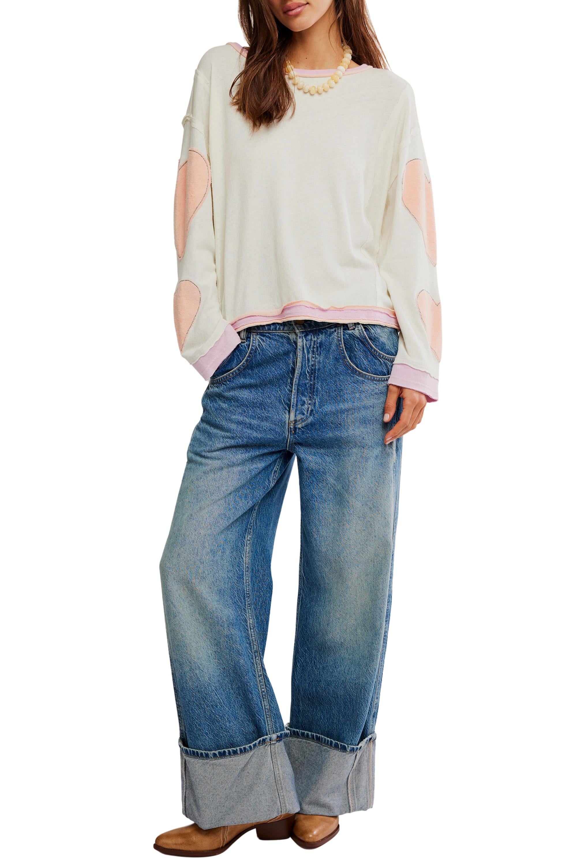 A woman wears the Free People Youre A Star Tee—an oversized white long-sleeve with pink star details—paired with wide-leg cuffed blue jeans, brown shoes, and a yellow beaded necklace, standing with one hand in her pocket.