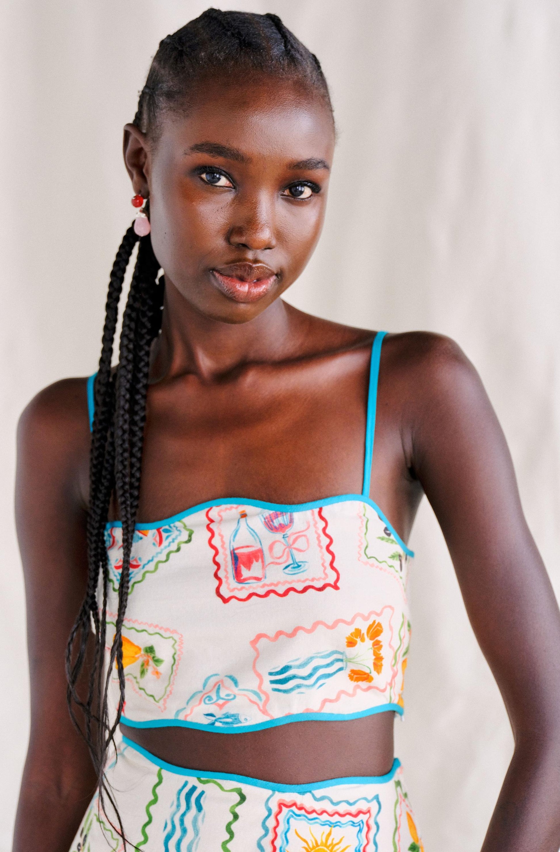 A woman with long braided hair models the Saylor Tashi Floral Set Top, a colorful, stamp-print linen crop top with thin blue straps. This imported piece from India highlights her style as she stands against a neutral background.