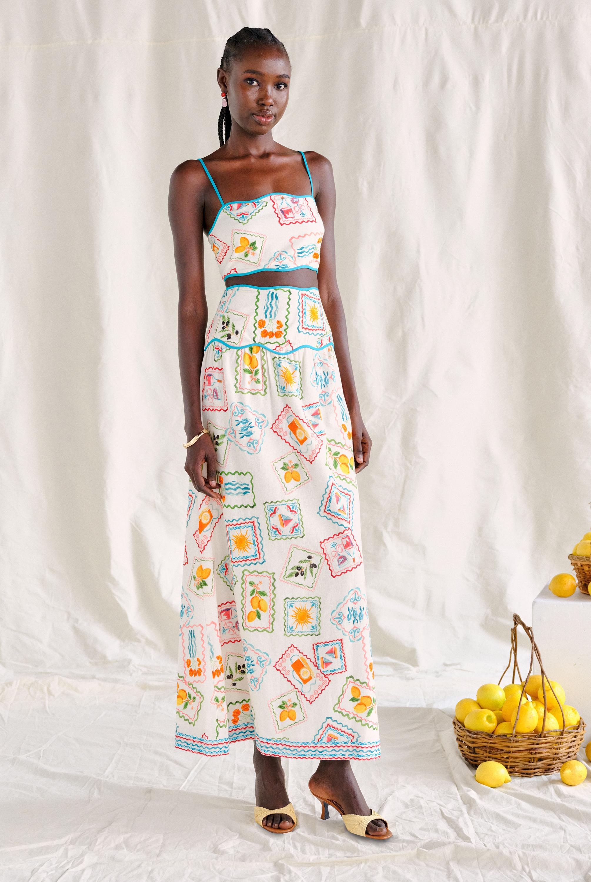 A woman with braided hair stands on a white backdrop wearing the Saylor Tashi Floral Set Top. She pairs it with sandals, and a basket of lemons sits beside her. The top features a colorful floral print and is imported from India.