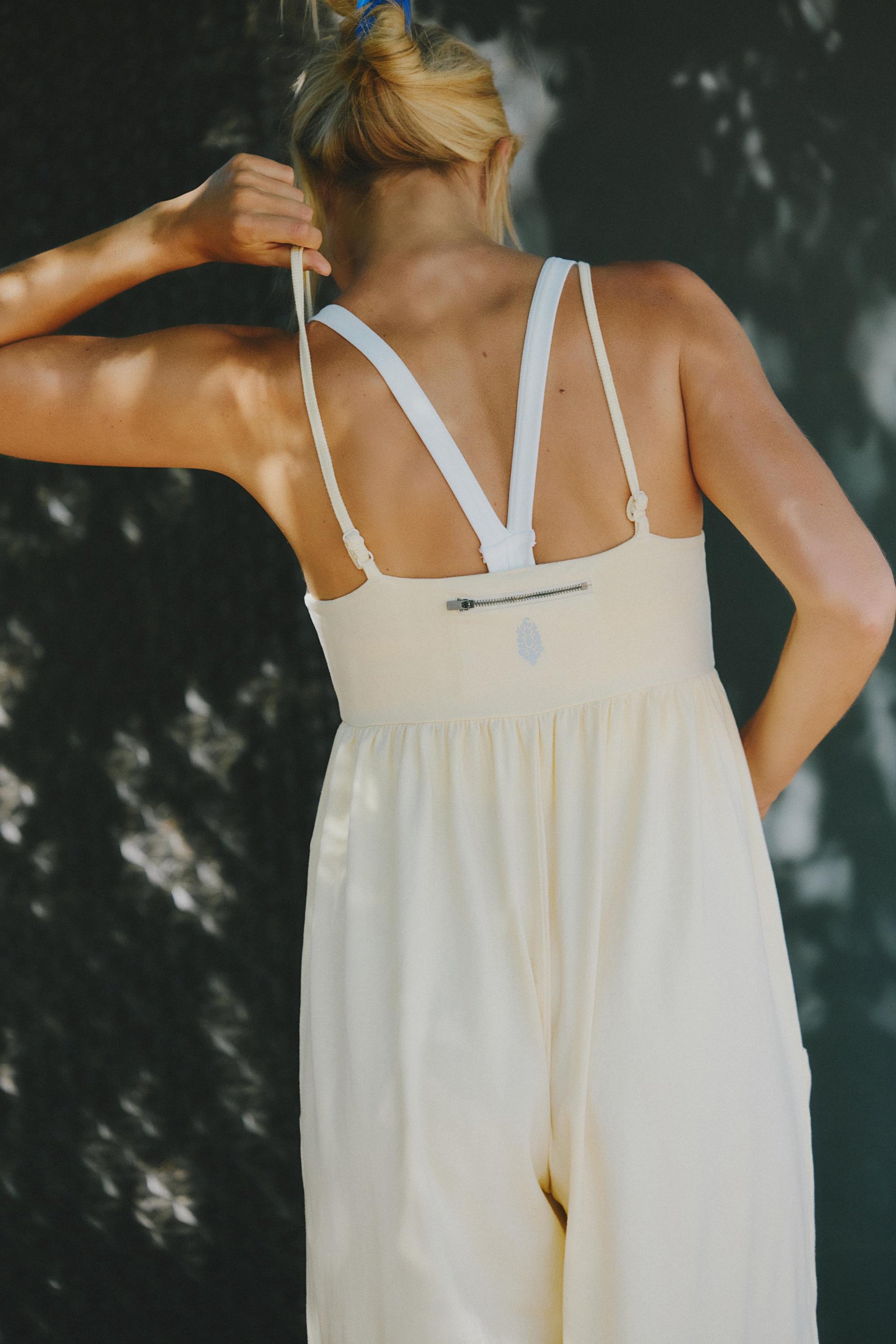 A blonde woman in a ponytail is seen from behind, adjusting the strap of the FP Movement Hot Shot Strappy Jumpsuit in Sunlit Yellow, featuring white straps and a babydoll-inspired design, against a dark background with dappled sunlight.