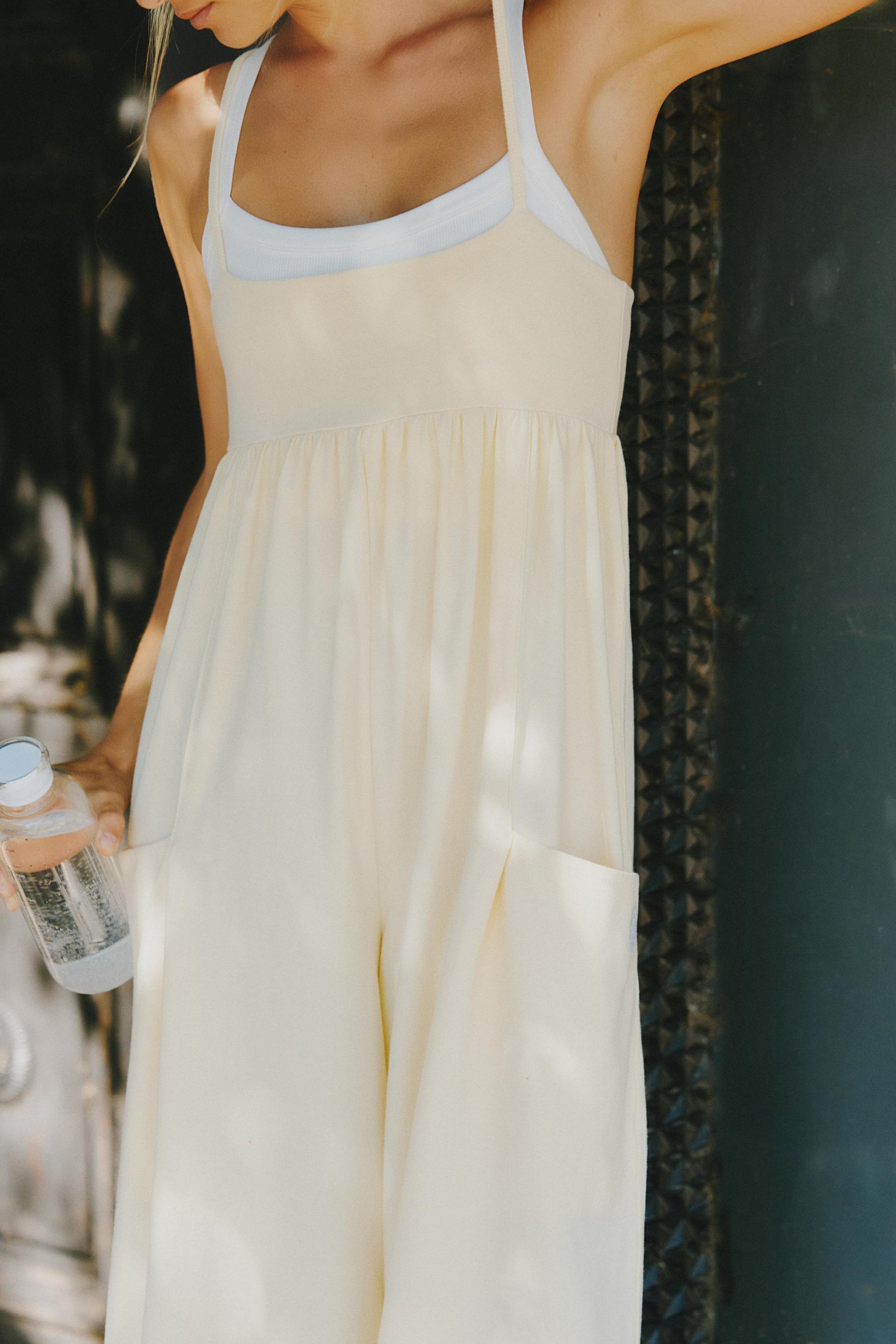 Someone stands outdoors in the sunlight wearing the FP Movement Hot Shot Strappy Jumpsuit in Sunlit Yellow, holding a bottle of water. Their head is out of frame and shadows play across the jumpsuit’s wide straps and square neckline.