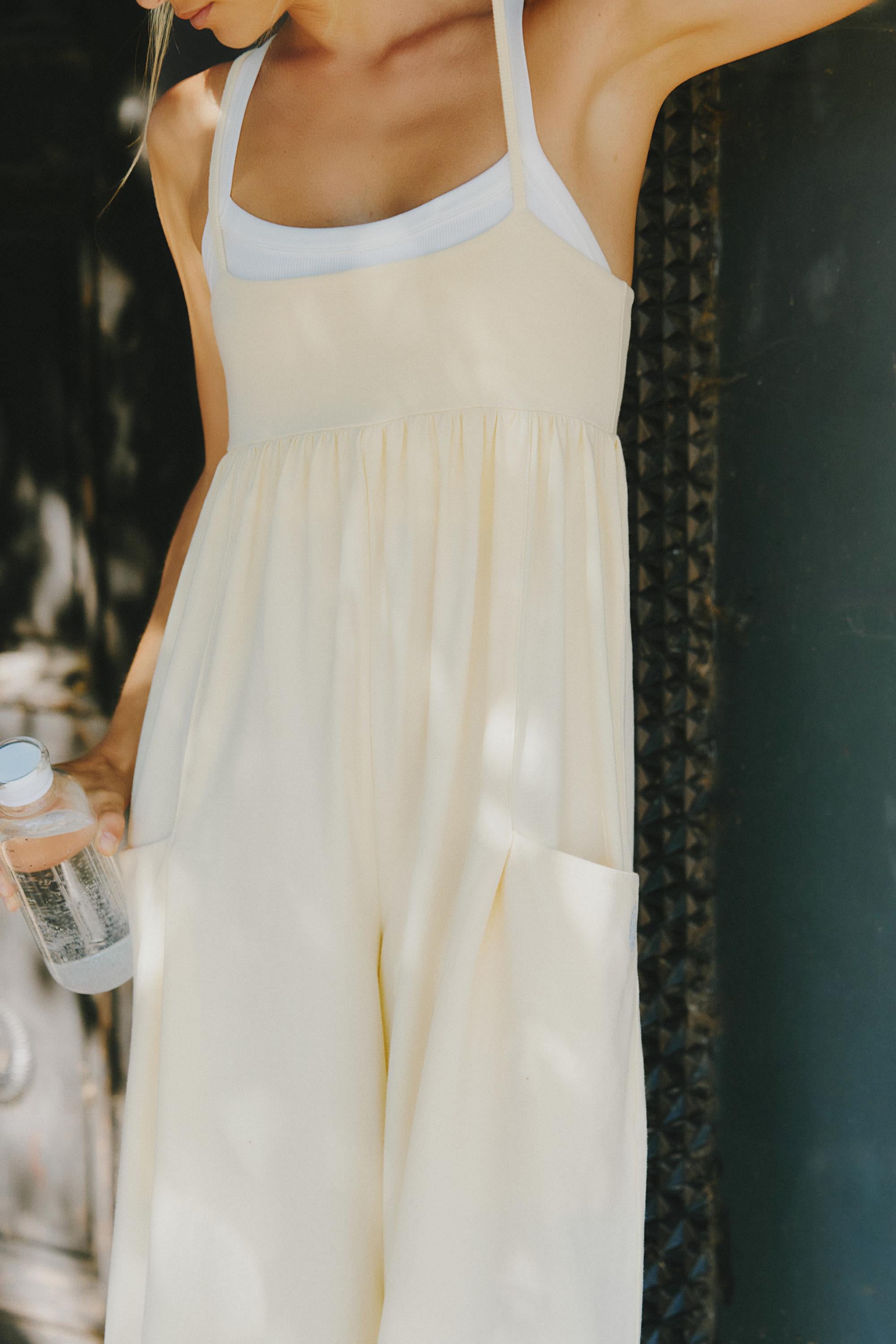Someone stands outdoors in the sunlight wearing the FP Movement Hot Shot Strappy Jumpsuit in Sunlit Yellow, holding a bottle of water. Their head is out of frame and shadows play across the jumpsuit’s wide straps and square neckline.