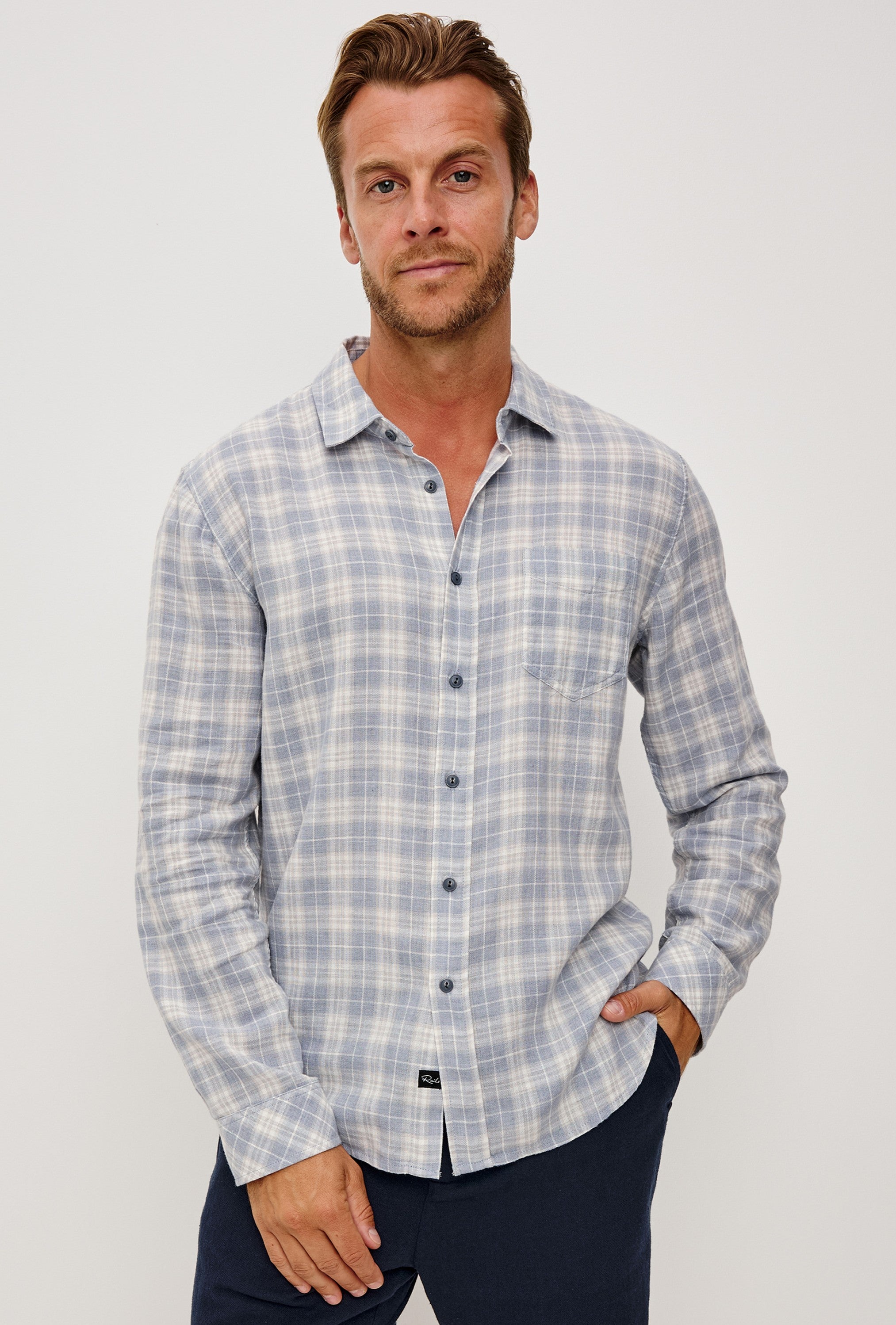 A man stands against a plain background, wearing the Rails Wyatt Thatch Cloud Cream Plaid Woven shirt in a relaxed fit with dark pants, one hand tucked in his pocket and looking directly at the camera with an easygoing expression.