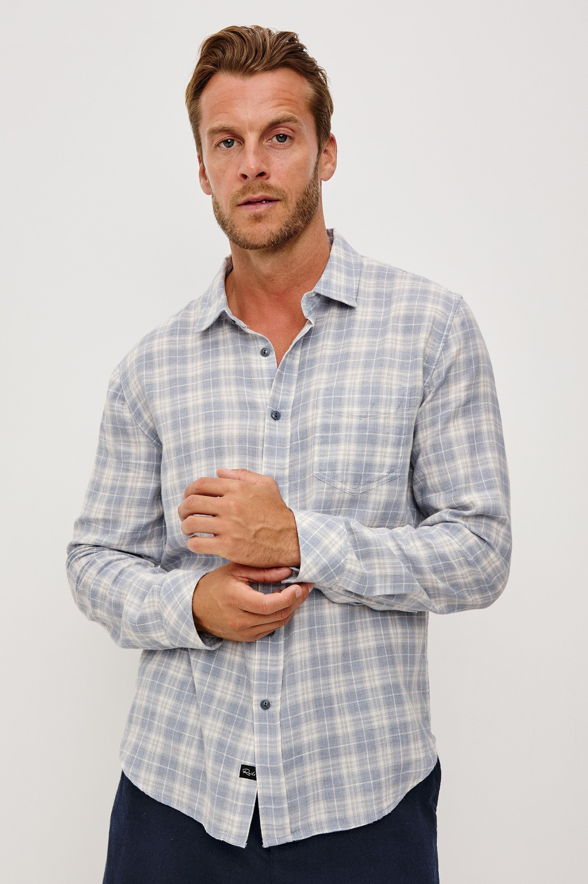A man with short hair and light stubble wears the Rails Wyatt Thatch Cloud Cream Plaid Woven shirt in light blue and white, standing against a plain light background with his hands loosely clasped in front of him.