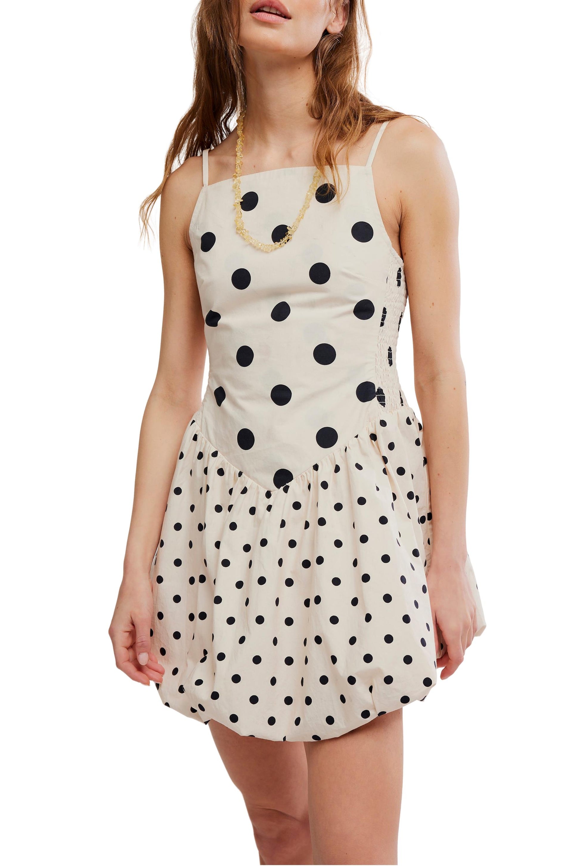 A woman wears the Misty Mini by Free People—a sleeveless, cream mini dress with black polka dots and a bubble hem. She pairs it with wavy hair and a gold necklace.