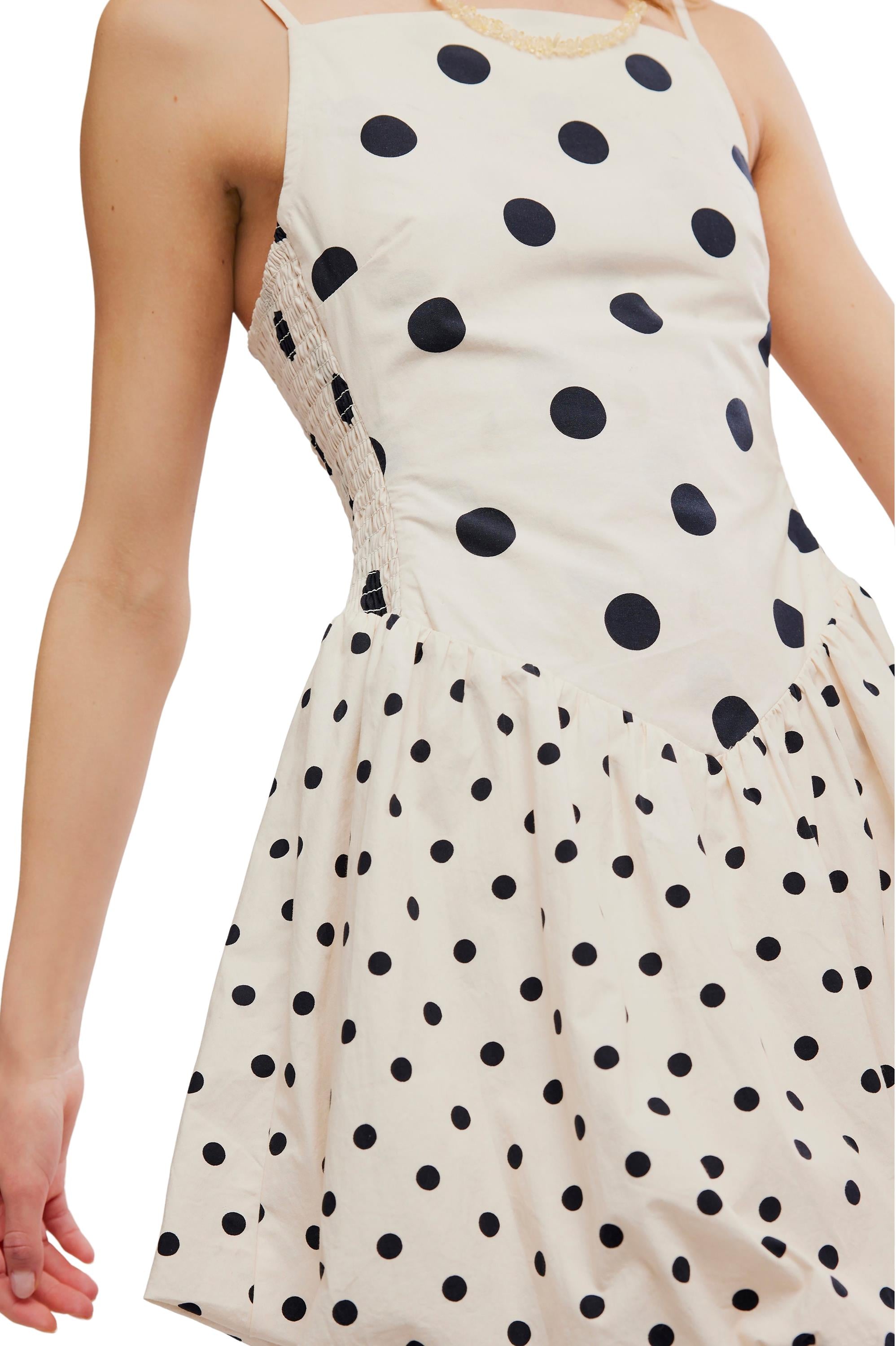 A person wears the Misty Mini by Free People, a sleeveless white dress with thin straps, black polka dots in various sizes, a fitted bodice, and a flared skirt.