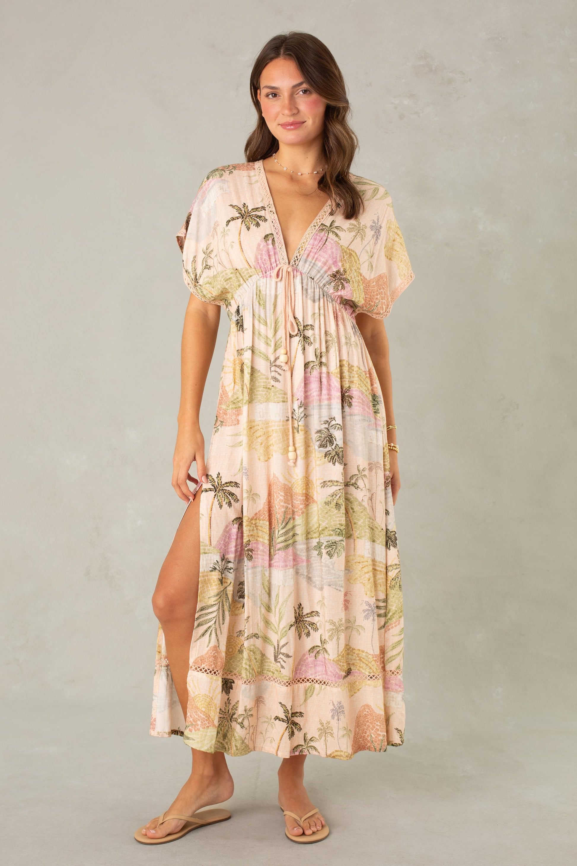 A woman models the Palma Maxi Coverup by South Moon Under, a light pastel maxi dress with a tropical landscape print, deep V-neckline, and short sleeves. She stands against a light gray background, holding one side of her dress and wearing sandals.
