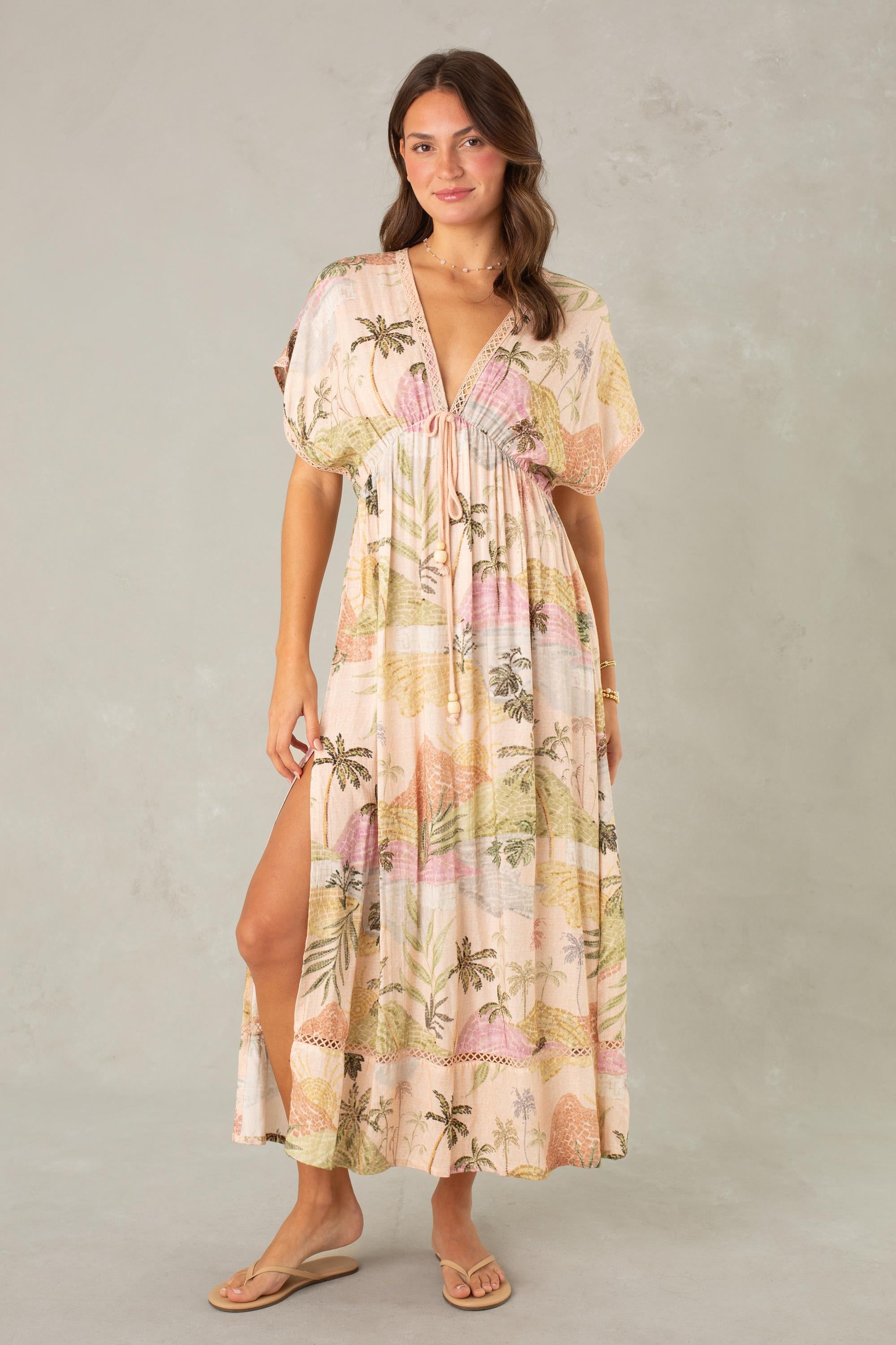 A woman models the Palma Maxi Coverup by South Moon Under, a light pastel maxi dress with a tropical landscape print, deep V-neckline, and short sleeves. She stands against a light gray background, holding one side of her dress and wearing sandals.