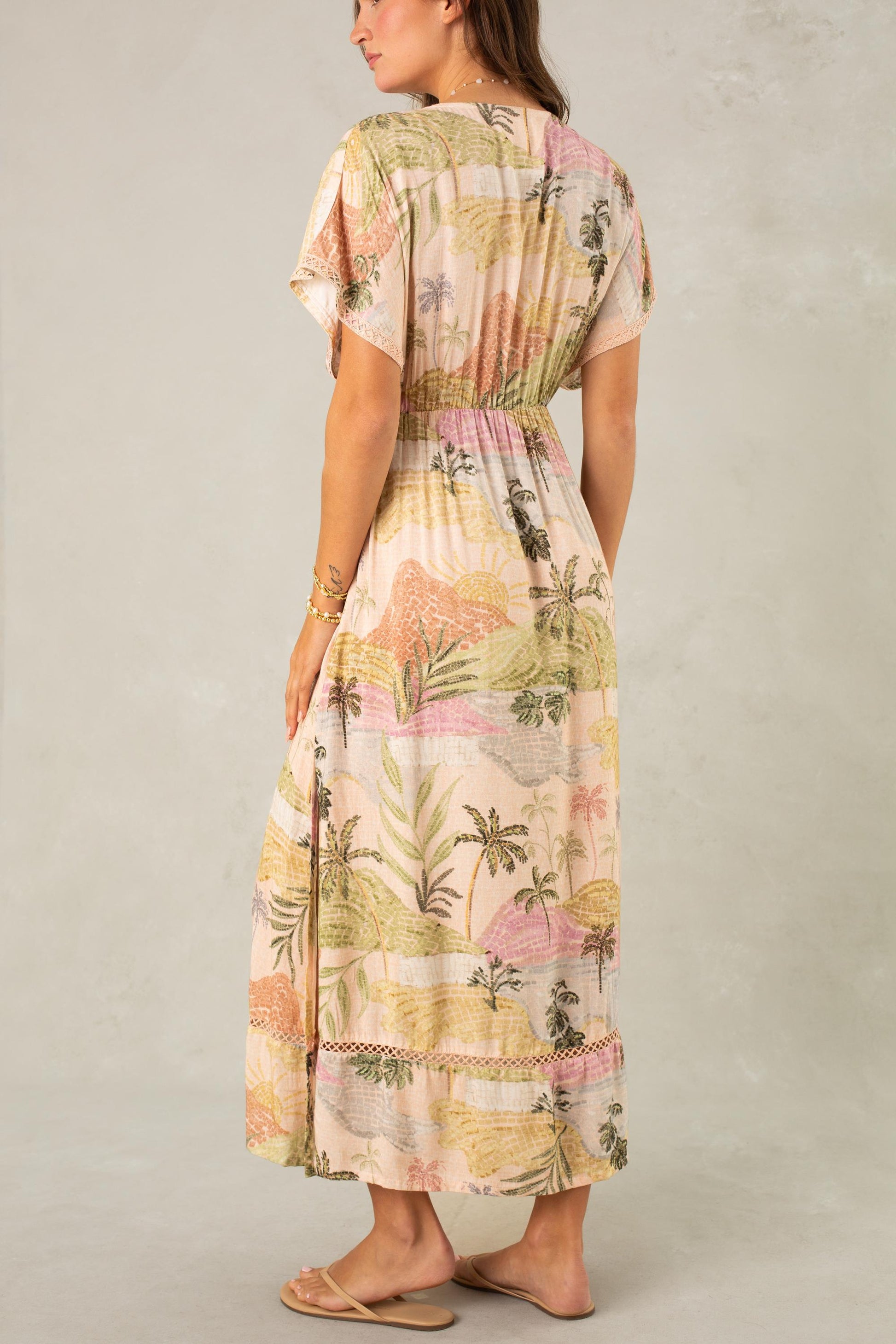 A woman, seen from behind against a light beige backdrop, wears the South Moon Under Palma Maxi Coverup—a short-sleeve, ankle-length, flowy maxi dress with a pastel tropical landscape print—paired with sandals.