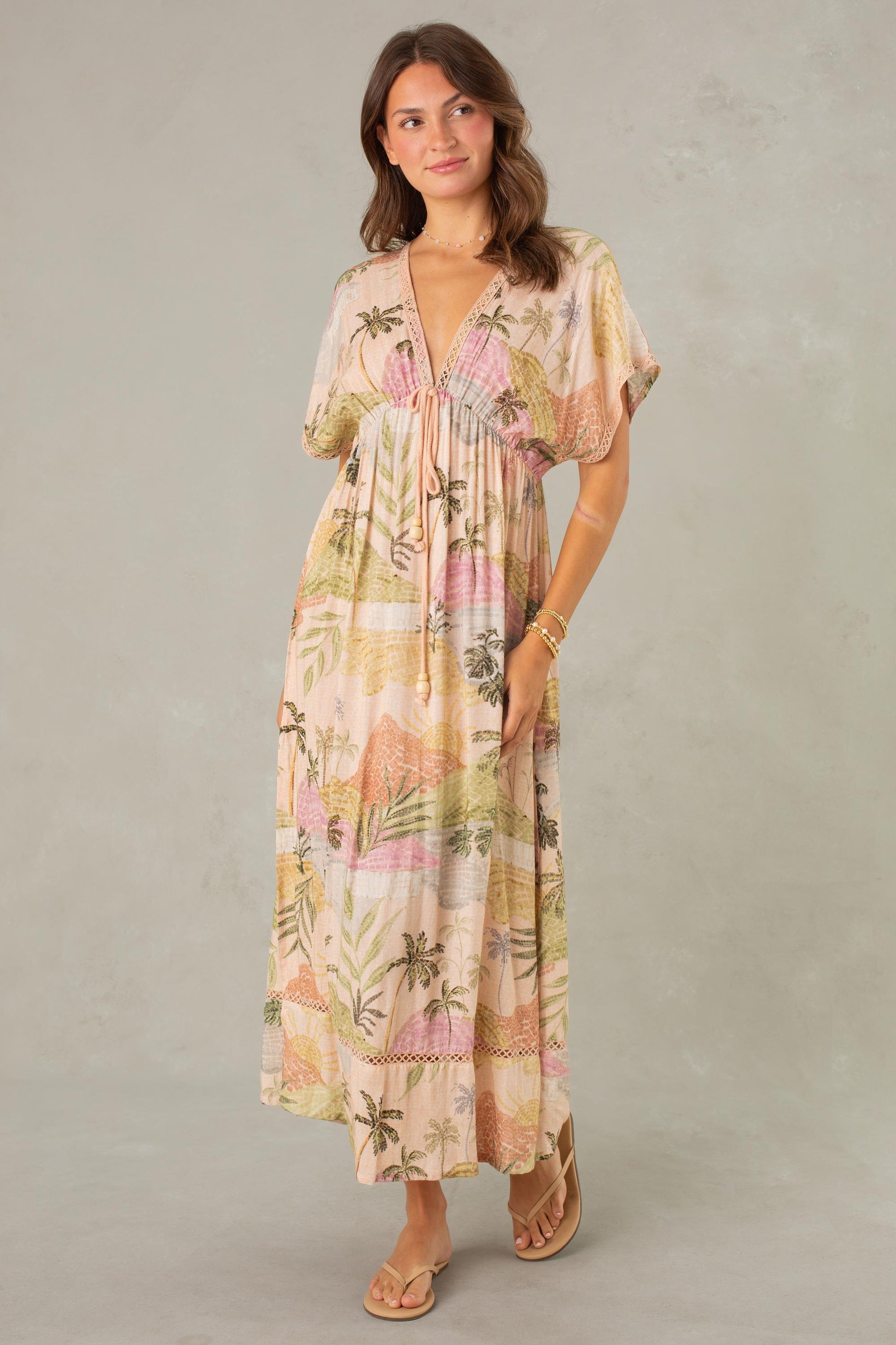 A woman wearing the South Moon Under Palma Maxi Coverup—a flowy, pastel-colored maxi dress with a tropical landscape print, short sleeves, and a soft deep V-neckline—stands against a light gray background, smiling gently and wearing sandals.