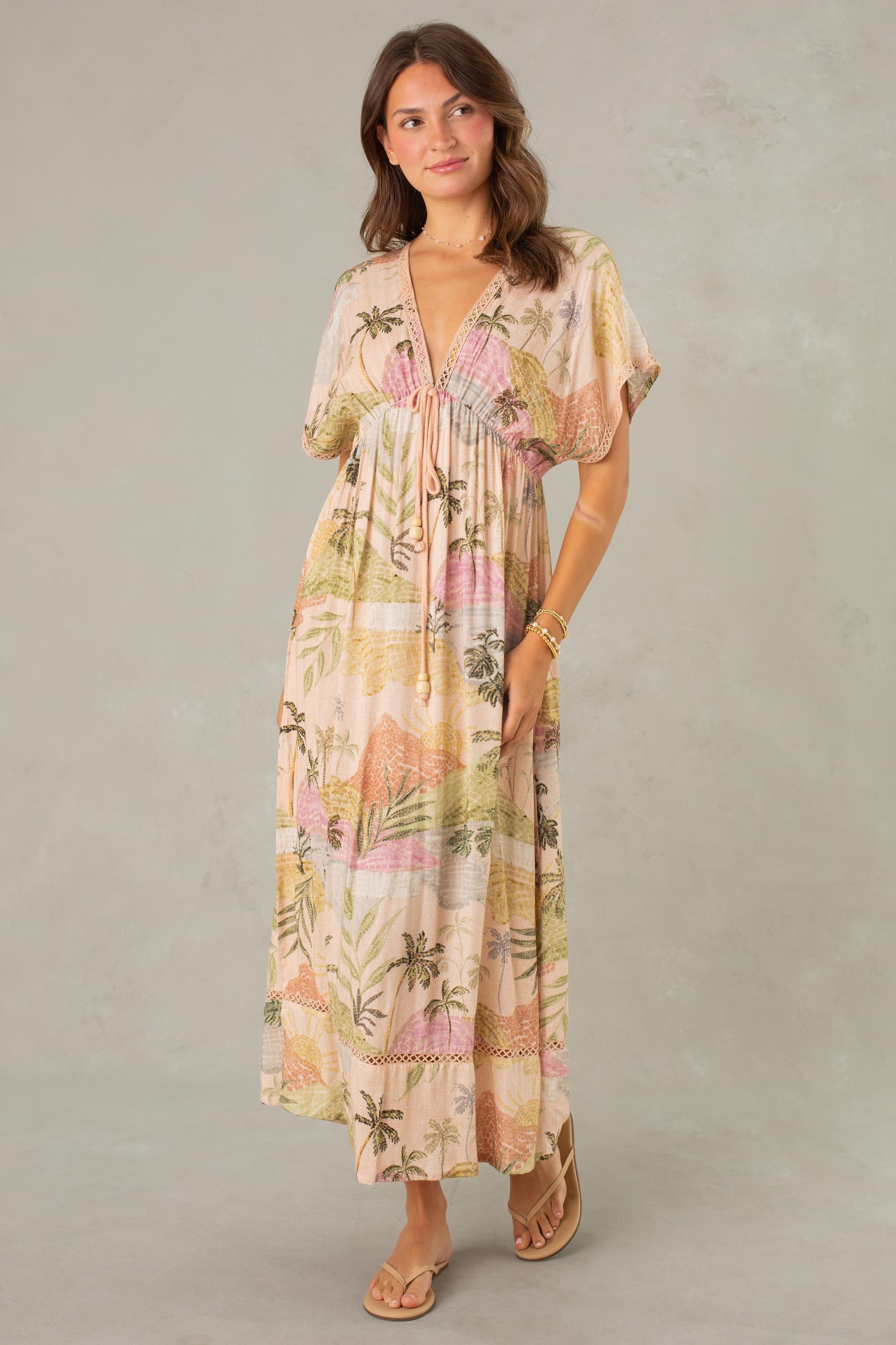 A woman wearing the South Moon Under Palma Maxi Coverup—a flowy, pastel-colored maxi dress with a tropical landscape print, short sleeves, and a soft deep V-neckline—stands against a light gray background, smiling gently and wearing sandals.
