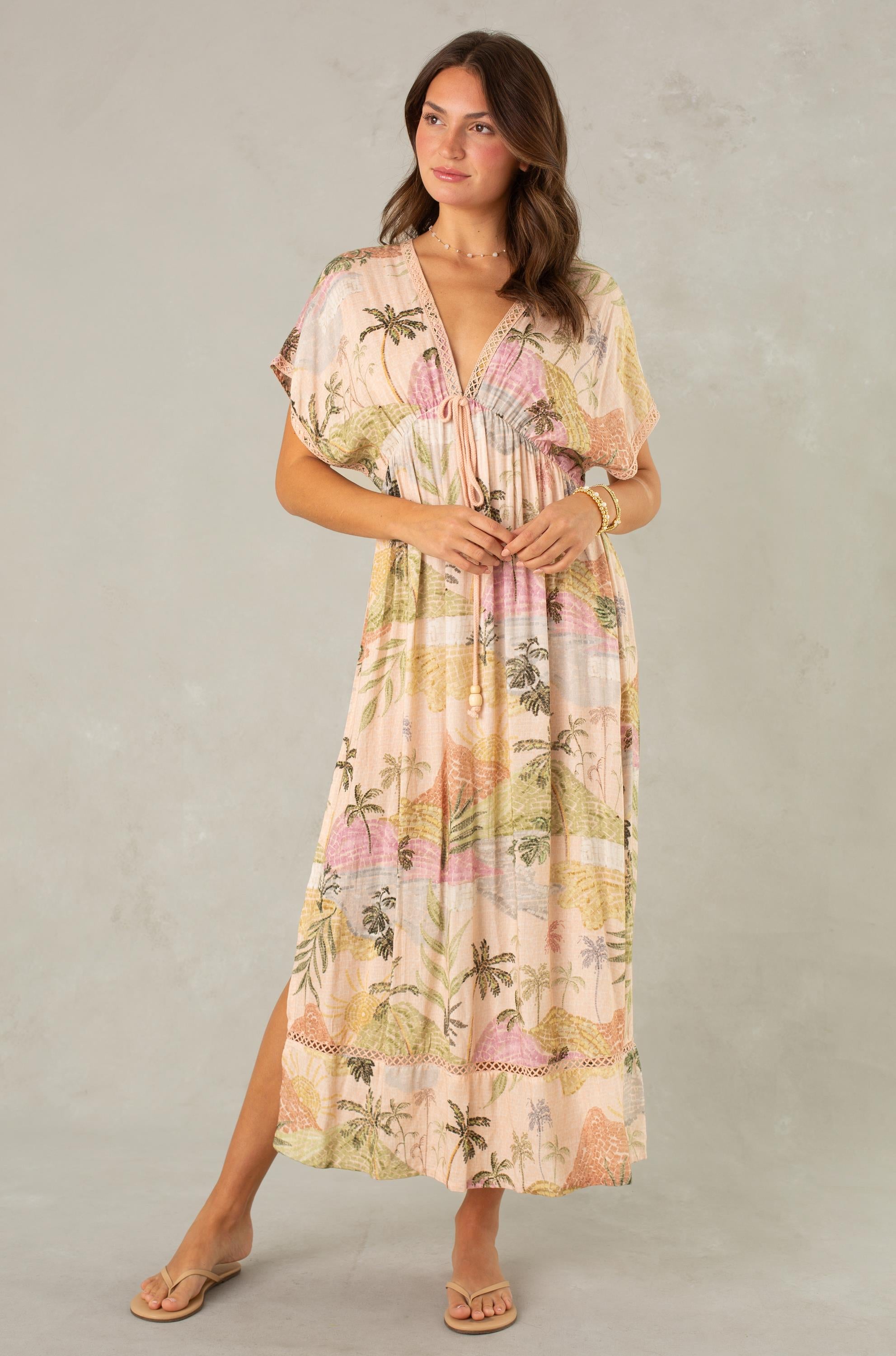 A woman models the South Moon Under Palma Maxi Coverup in MULTI, featuring a flowy silhouette with a tropical landscape print in soft, pastel hues. She stands against a light gray background, glancing to the side while holding the dress with one hand and resting her other hand on her waist.