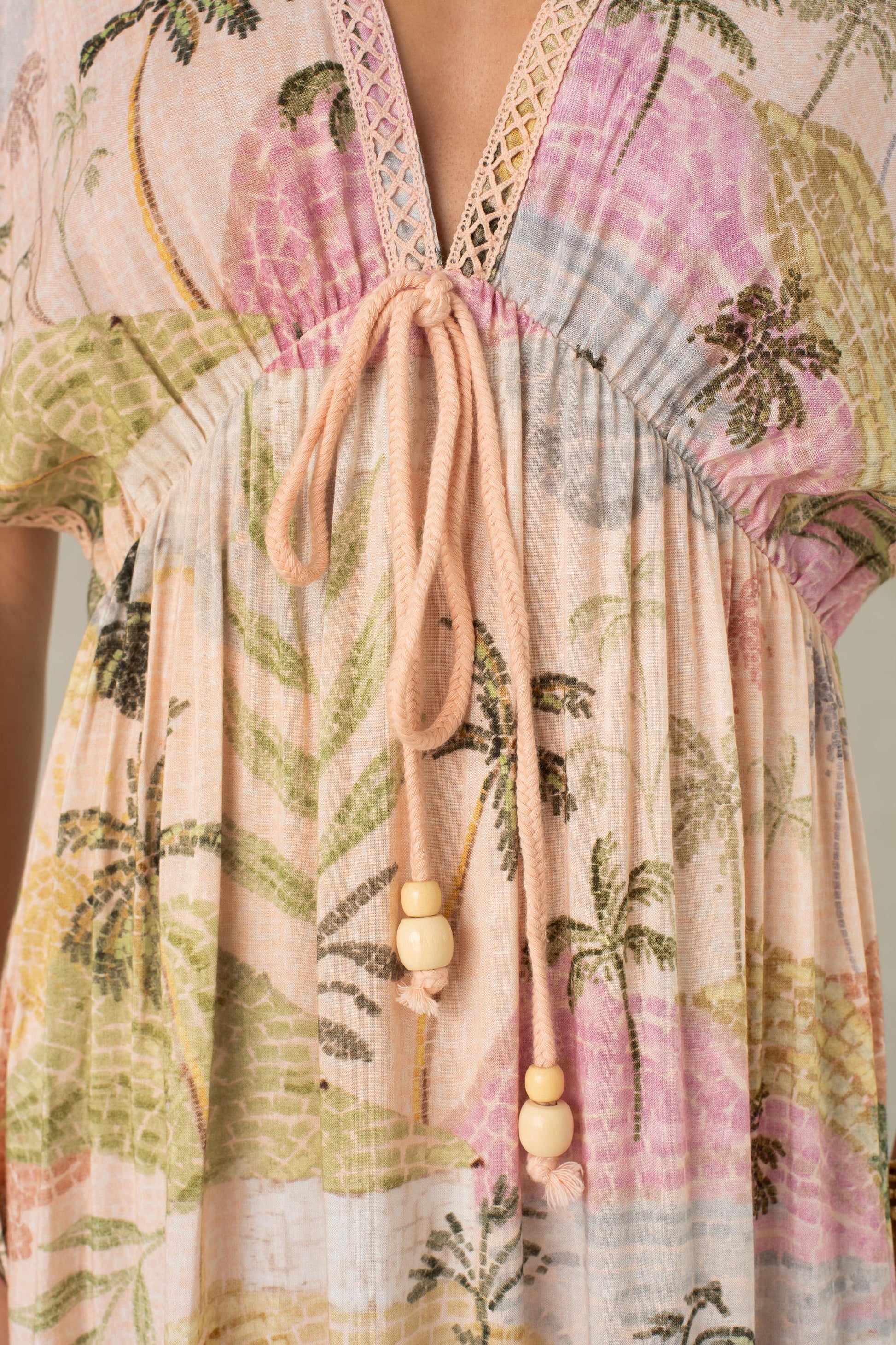 Close-up of the South Moon Under Palma Maxi Coverup—a light, flowy maxi dress showcasing a pastel tropical landscape print, designed with a V-neckline and adjustable pink rope drawstring ties with wooden bead accents.
