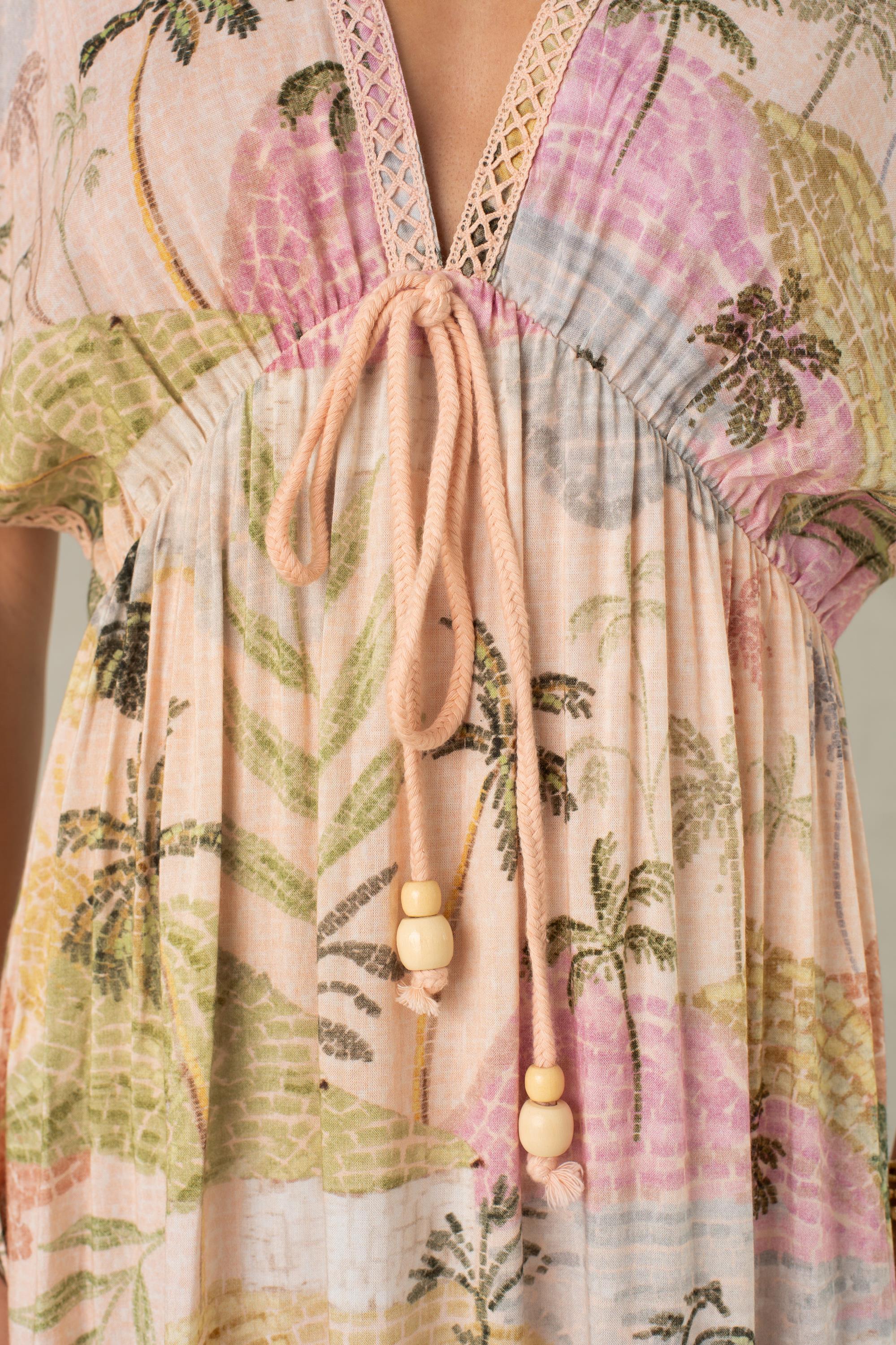 Close-up of the South Moon Under Palma Maxi Coverup—a light, flowy maxi dress showcasing a pastel tropical landscape print, designed with a V-neckline and adjustable pink rope drawstring ties with wooden bead accents.