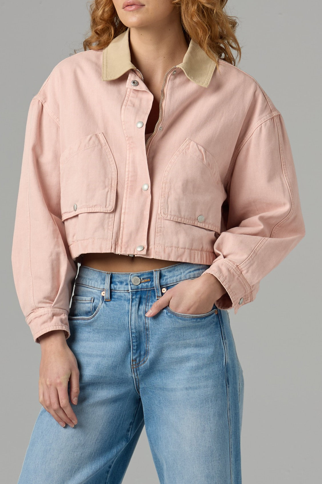 A woman with wavy hair wears the Blank Lawn Party Barn Jacket in LAWN PARTY - PINK COMBO, styled with blue jeans. She stands against a plain gray background, one hand on her hip, highlighting the jacket’s relaxed, utility-inspired fit and large pockets.