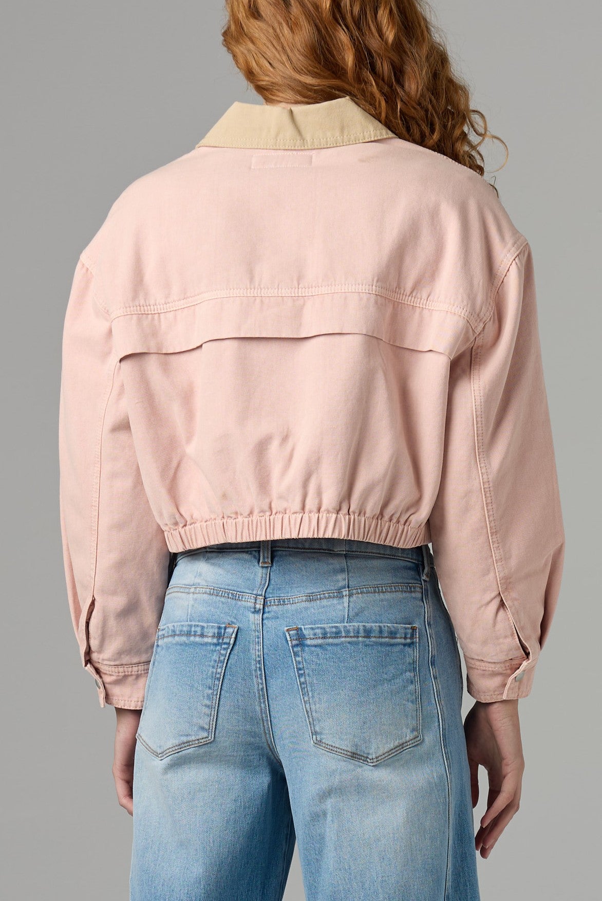 A person with wavy red hair is shown from the back wearing the Blank Lawn Party Barn Jacket—a light pink, utility-inspired collared jacket with an elastic waistband—paired with light blue jeans.