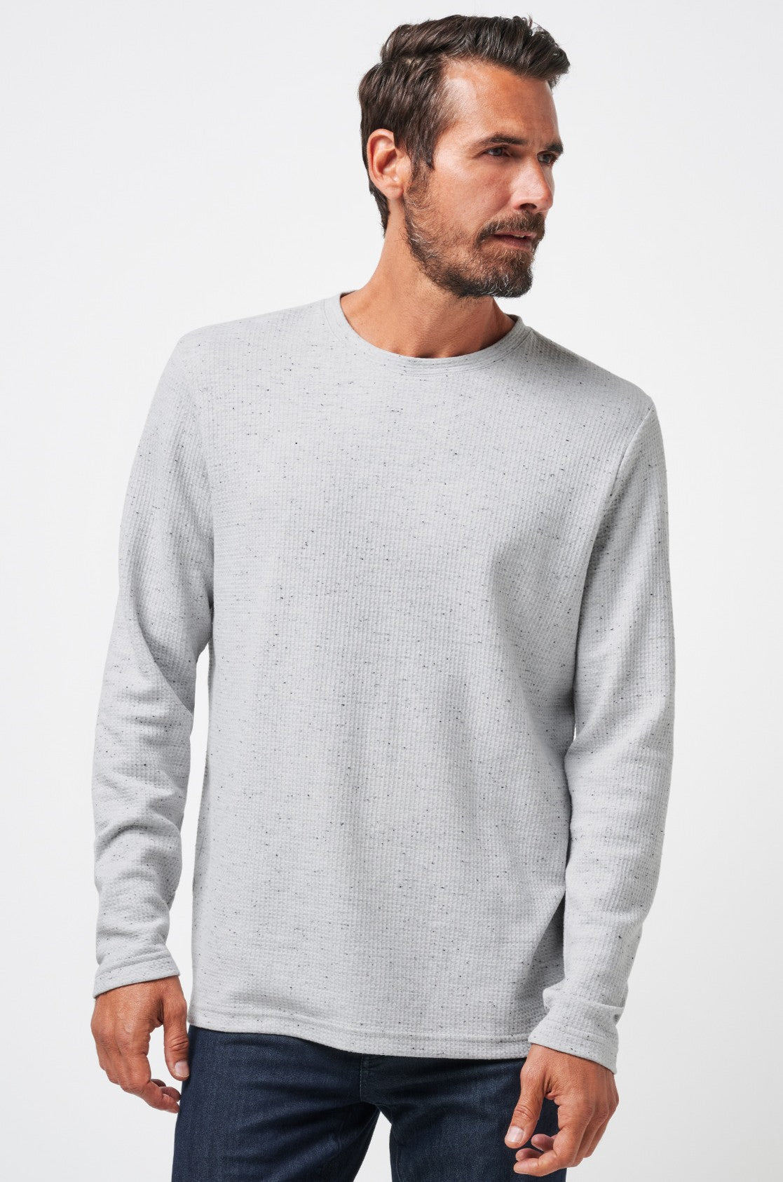 A man with short dark hair and a beard wears the TravisMathew Cloud Waffle Crew in light grey, paired with dark jeans. He stands against a plain white background, gazing slightly to his right.