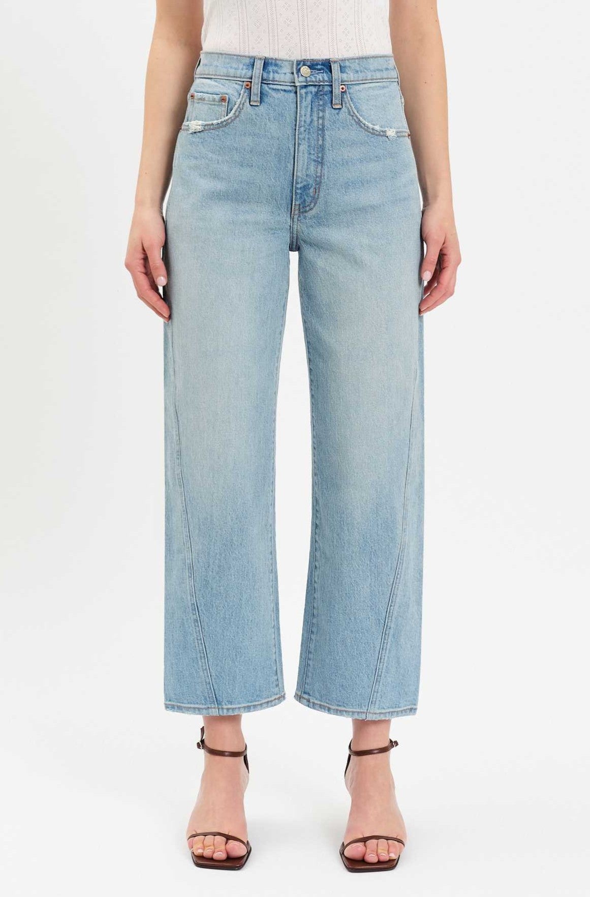 A woman stands facing forward, wearing the Daze Westward Curved Straight Denim in PULSE—a high-rise, wide-leg silhouette with a cropped ankle length. She pairs the jeans with black heeled sandals and a white sleeveless top against a plain white background.