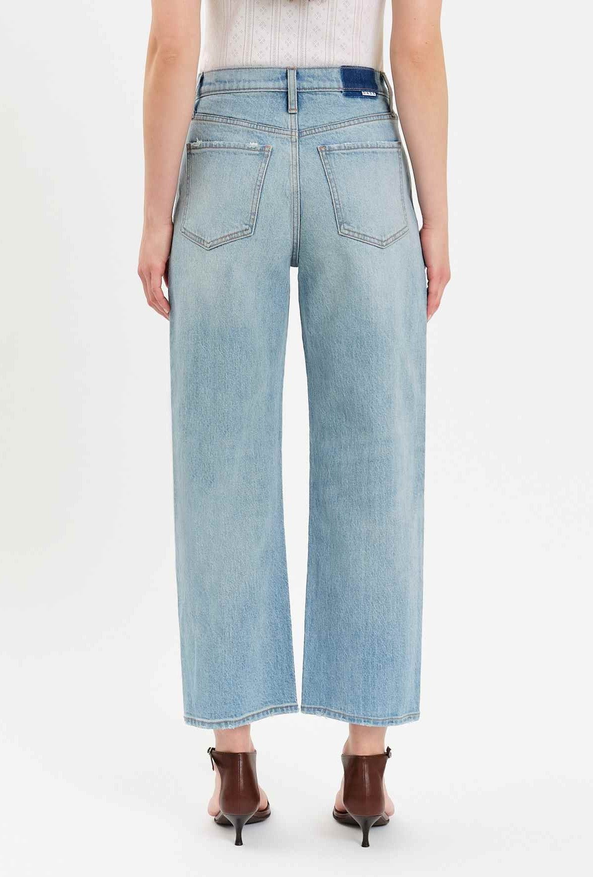 A person is shown from the back wearing Daze’s Westward Curved Straight Denim jeans in light blue. The high-rise, straight-leg jeans feature two back pockets and hit above the ankles, paired with heeled sandals and a sleeveless white top.