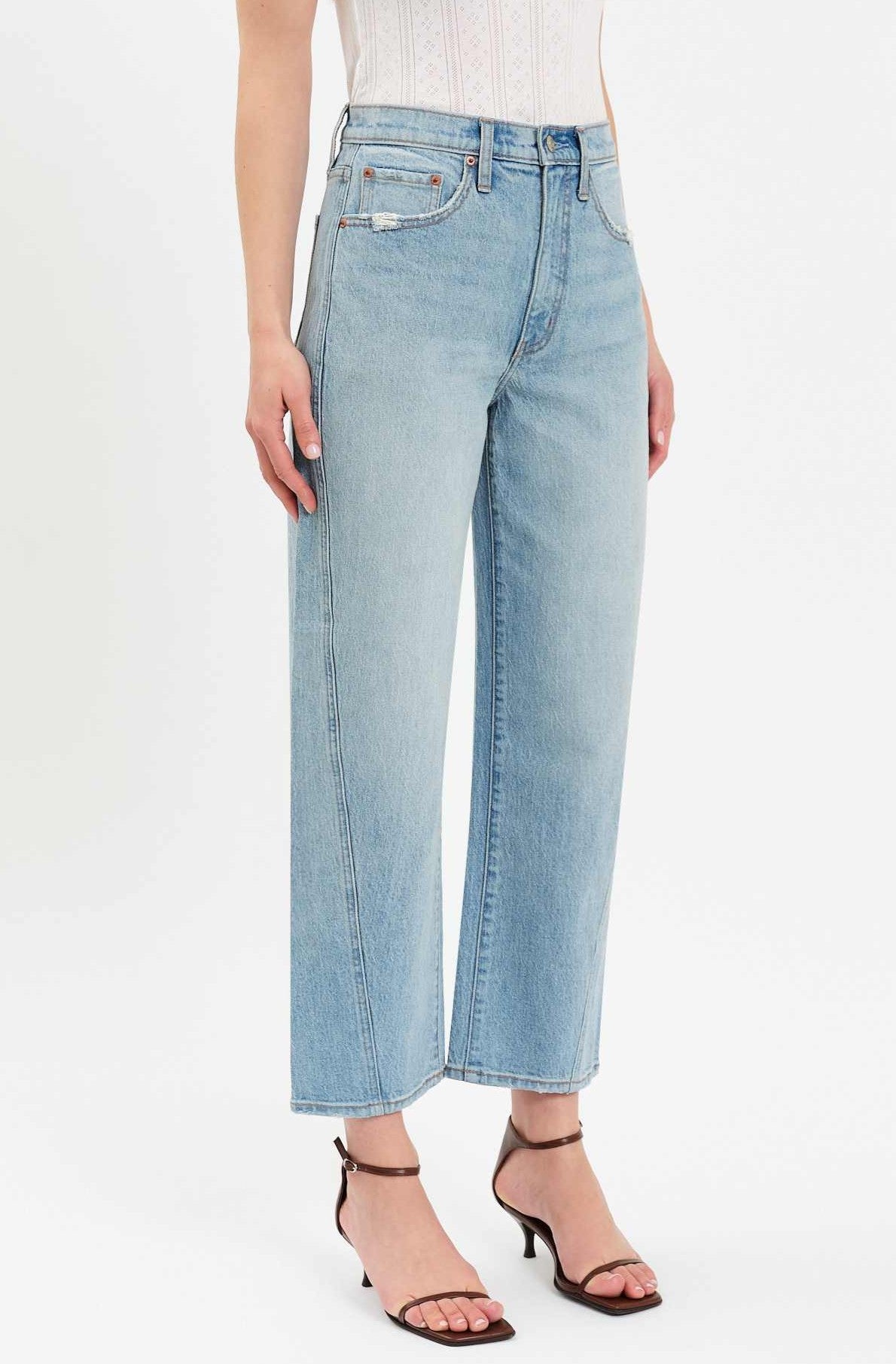 A person wearing the Daze Westward Curved Straight Denim in light blue, styled with a white sleeveless top and black heeled sandals, stands against a plain white background.