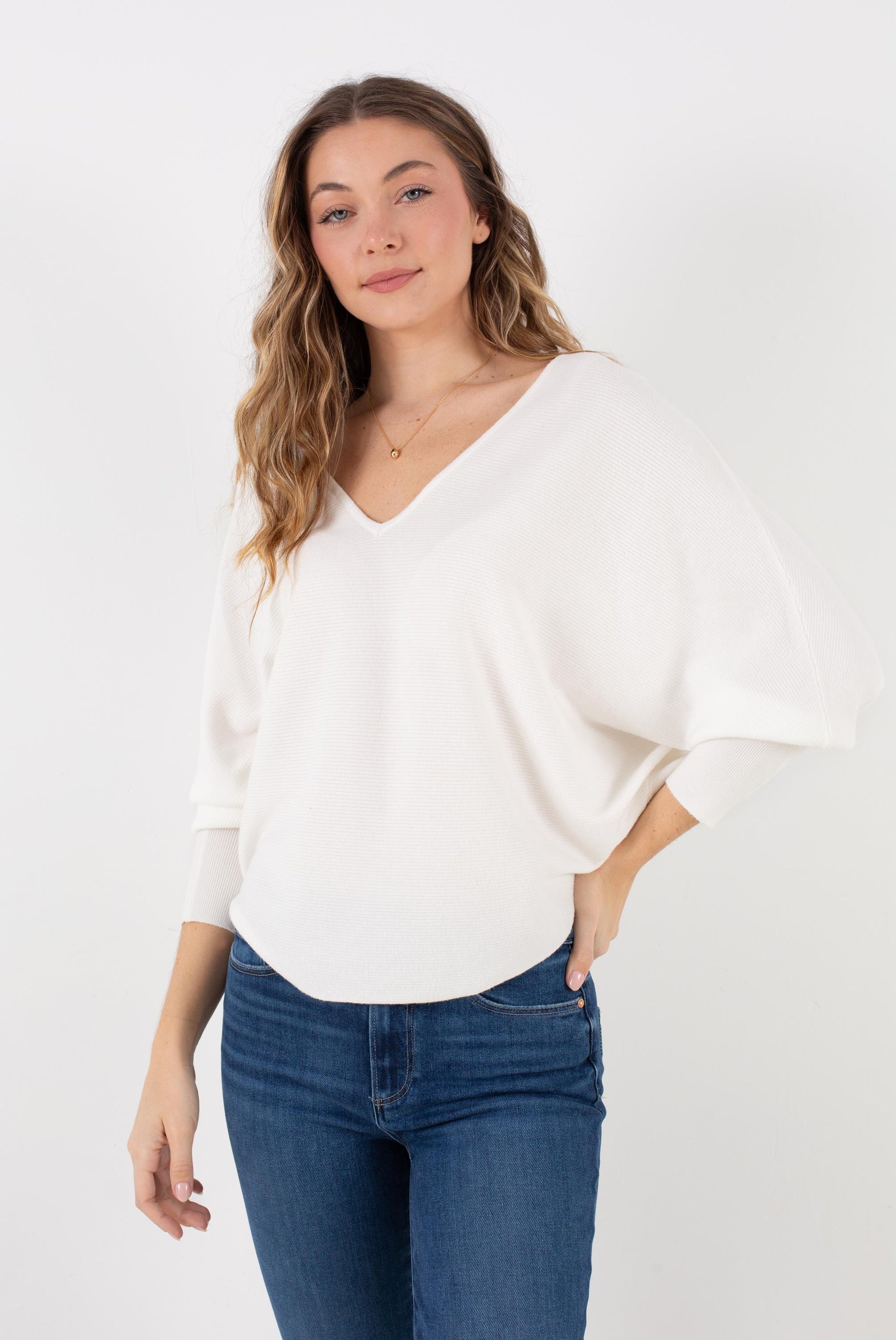 A woman with wavy hair wears the Kerisma Dolman Ryu V Neck in Ivory, paired with blue jeans, standing against a plain white background with a relaxed, confident expression.