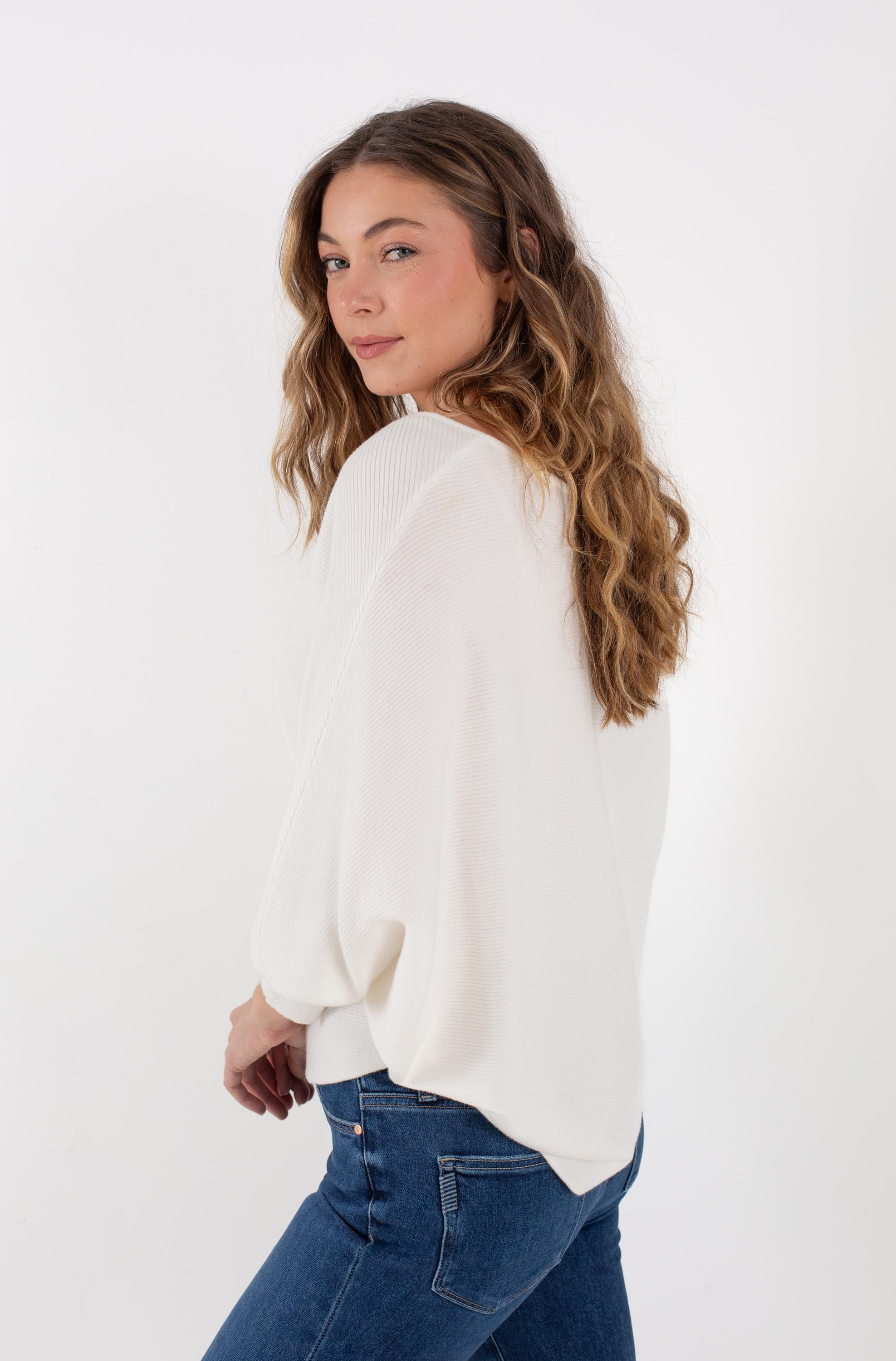 A woman with long wavy hair is wearing the Kerisma Dolman Ryu V Neck in Ivory, styled with blue jeans, standing sideways and looking over her shoulder against a plain white background.
