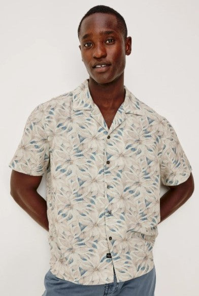 A man smiles slightly with his hands behind his back, wearing the Rails Rhodes shirt—a vintage-inspired, short-sleeve button-up featuring a beige and blue floral pattern—standing against a plain background.