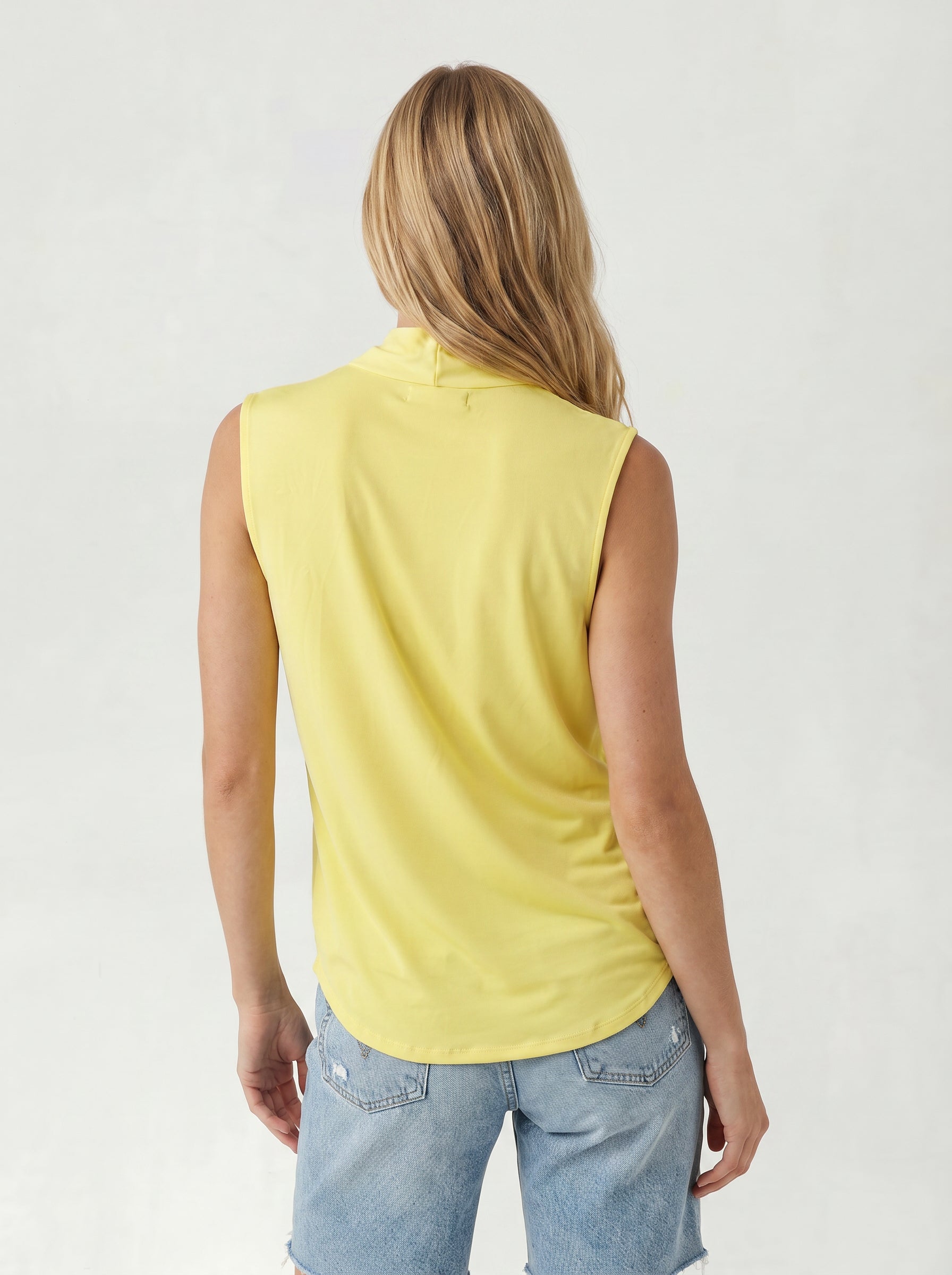 A person with long blonde hair is seen from behind wearing the Georgie Gwen Sleeveless Wrap Top in yellow and light blue denim shorts against a plain light background.