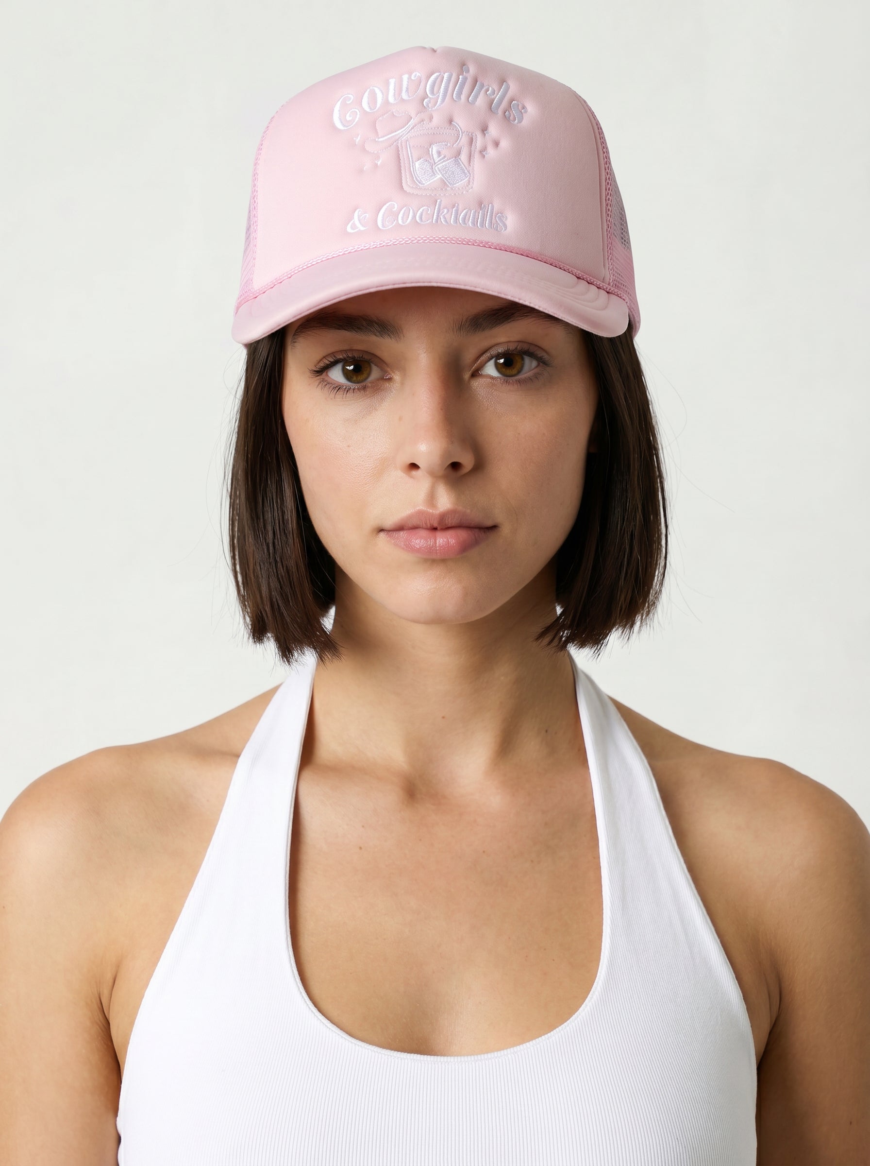 A woman with straight dark hair wears the South Moon Under Cowgirls & Cocktails Trucker Hat in light pink and a white halter top, standing against a plain light background.
