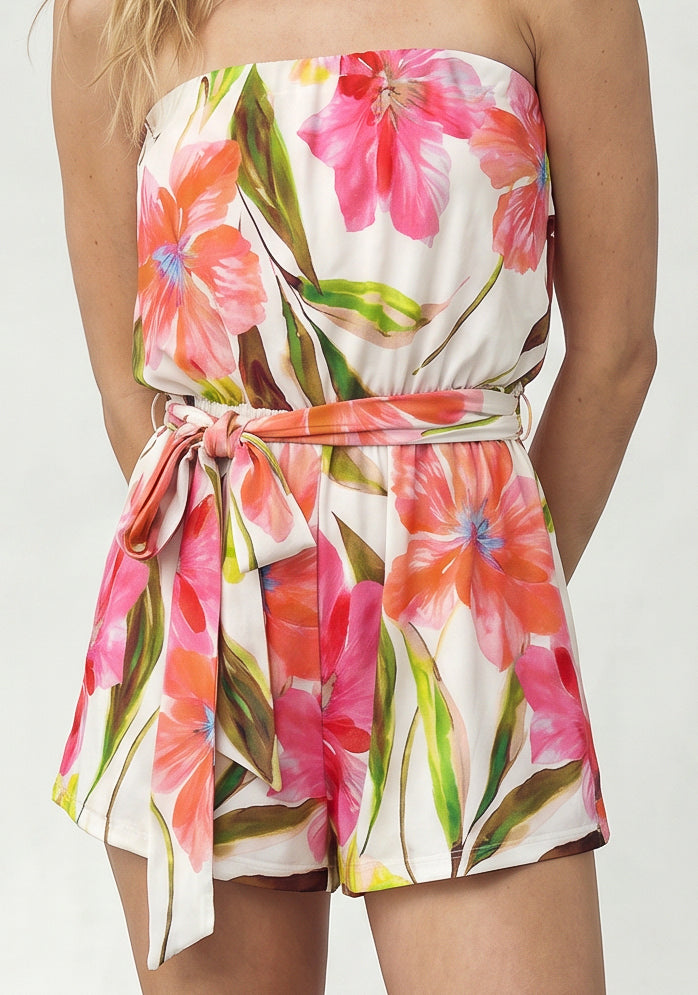 A woman wears the Isla Ari Strapless Easy Romper in White Multi, featuring a vibrant floral print, flowy shorts, and a matching fabric belt tied at the waist.