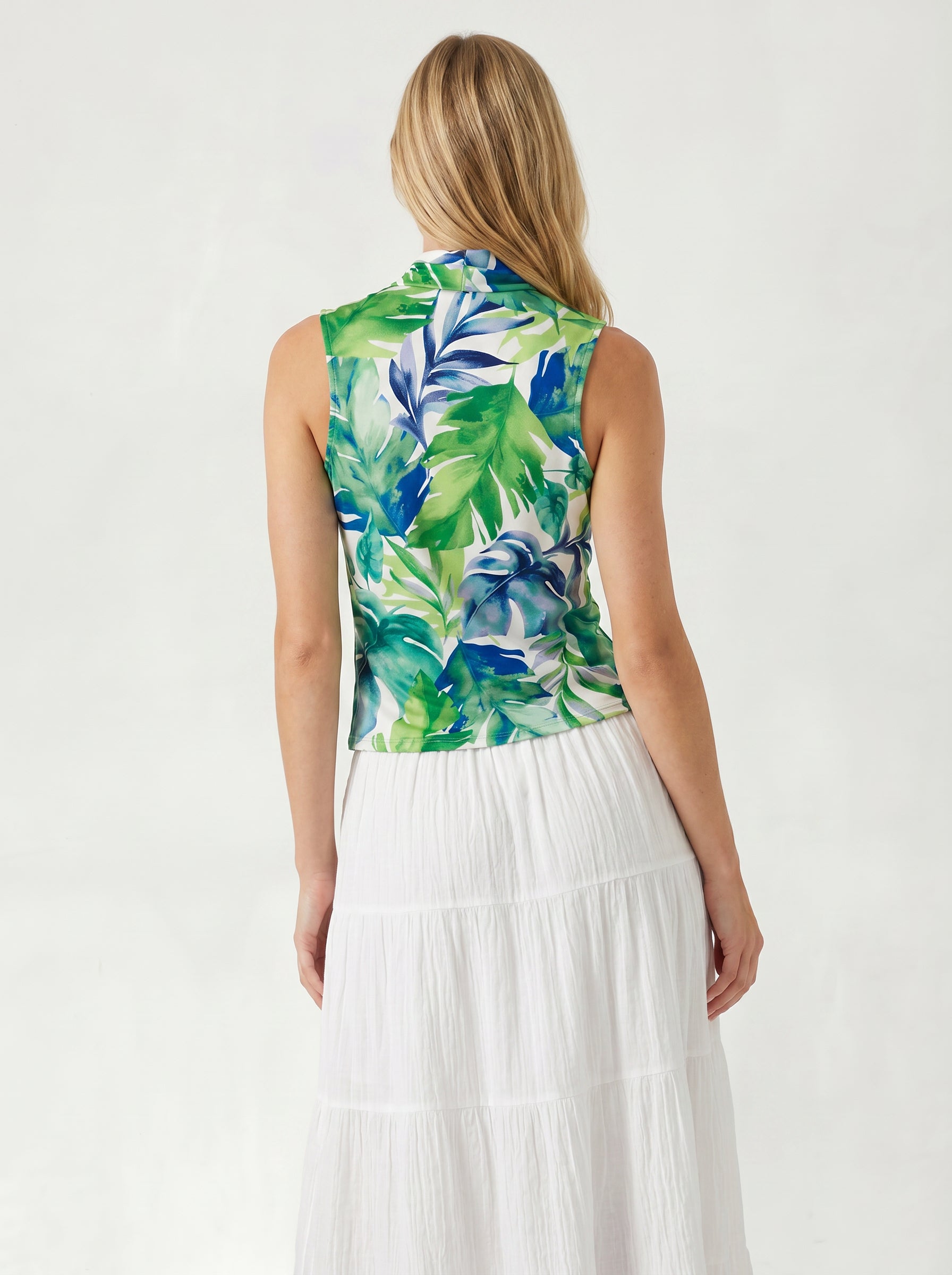 A woman with blonde hair, seen from behind, wears the Georgie Gwen Sleeveless Wrap Top featuring a green and blue tropical leaf print, paired with a long white tiered skirt. The light background highlights her resort-inspired style.