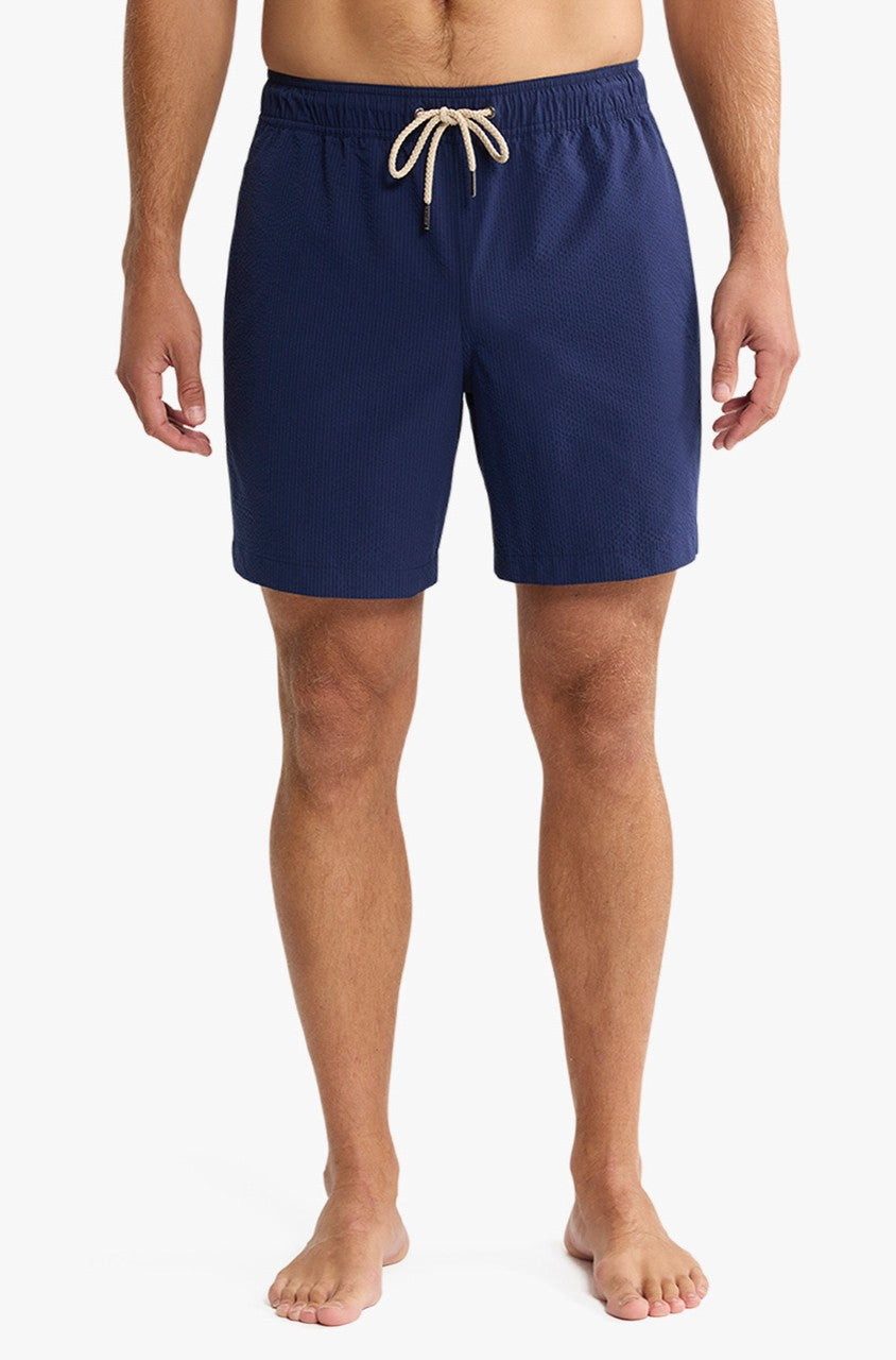 A barefoot person stands against a white background, wearing Fair Harbor's Bayberry Trunk 7 Inch Navy Seersucker shorts. The image is cropped at the shoulders, emphasizing the shorts and legs.