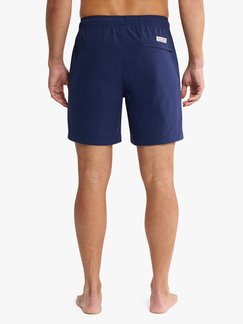 A man stands barefoot on a white background, shown from the back, wearing Fair Harbor's Bayberry Trunk 7 Inch Navy Seersucker shorts with an elastic waistband and a single right back pocket.