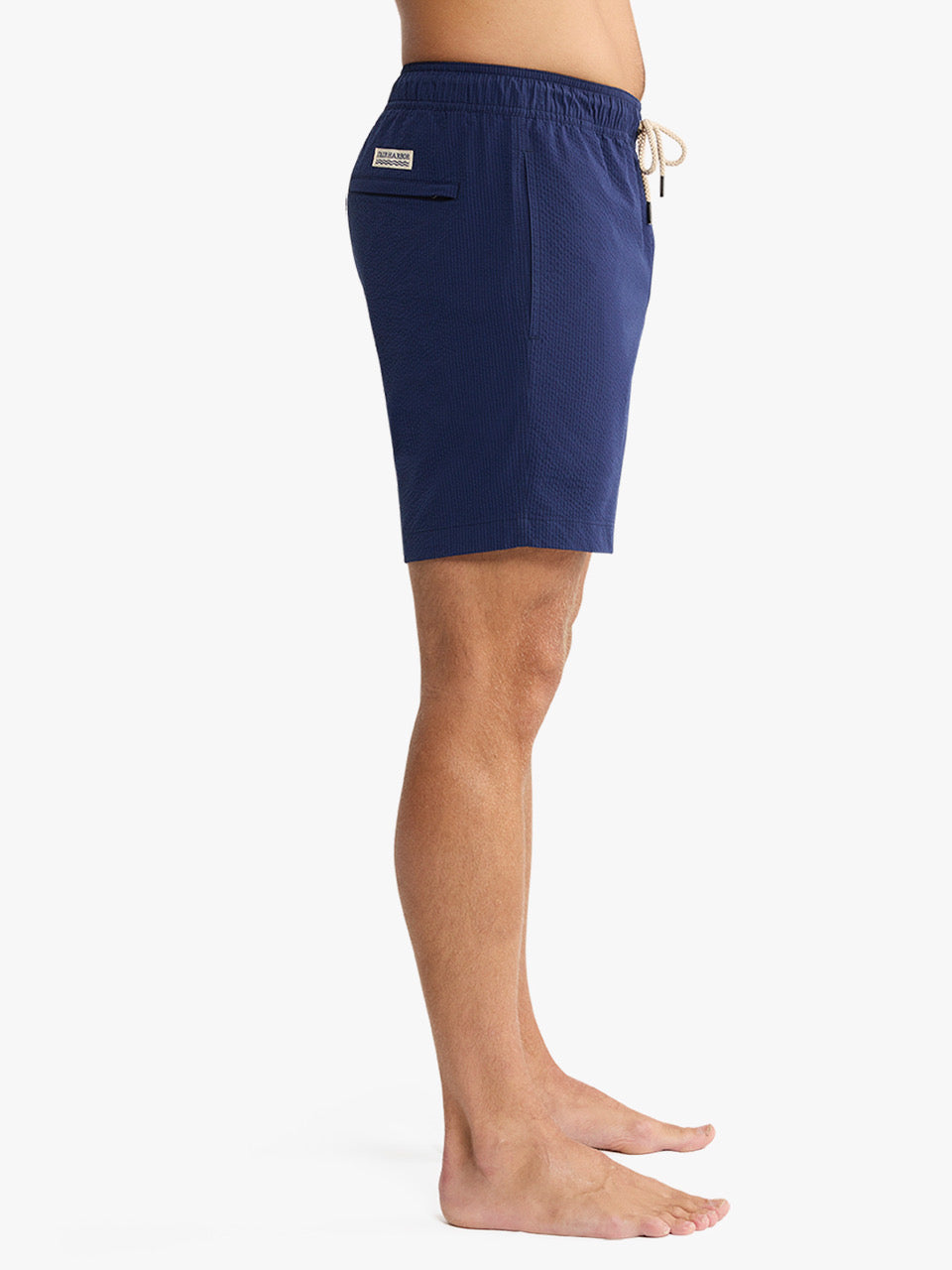 A person stands sideways against a white background wearing Fair Harbor’s Bayberry Trunk 7 Inch Navy Seersucker shorts, featuring a drawstring waist, built-in liner, back pocket, and a small label.