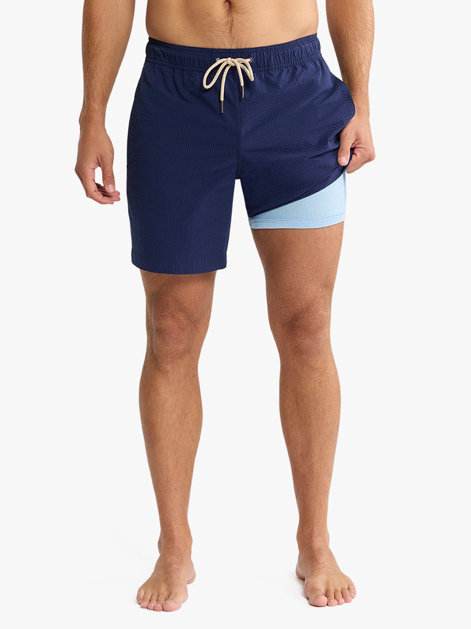 A man stands barefoot against a white background wearing Fair Harbor Bayberry Trunk 7 Inch Navy Seersucker shorts with a classic fit, light blue color block on the right leg, built-in liner, and beige drawstring at the waist.