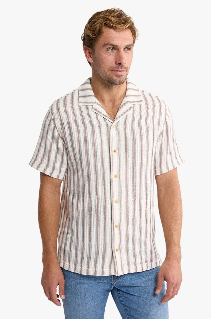 A man wears the Fair Harbor Waveloom Crochet Shirt, a relaxed fit, short-sleeve button-up with vertical beige and white stripes, paired with light blue jeans against a plain white background.