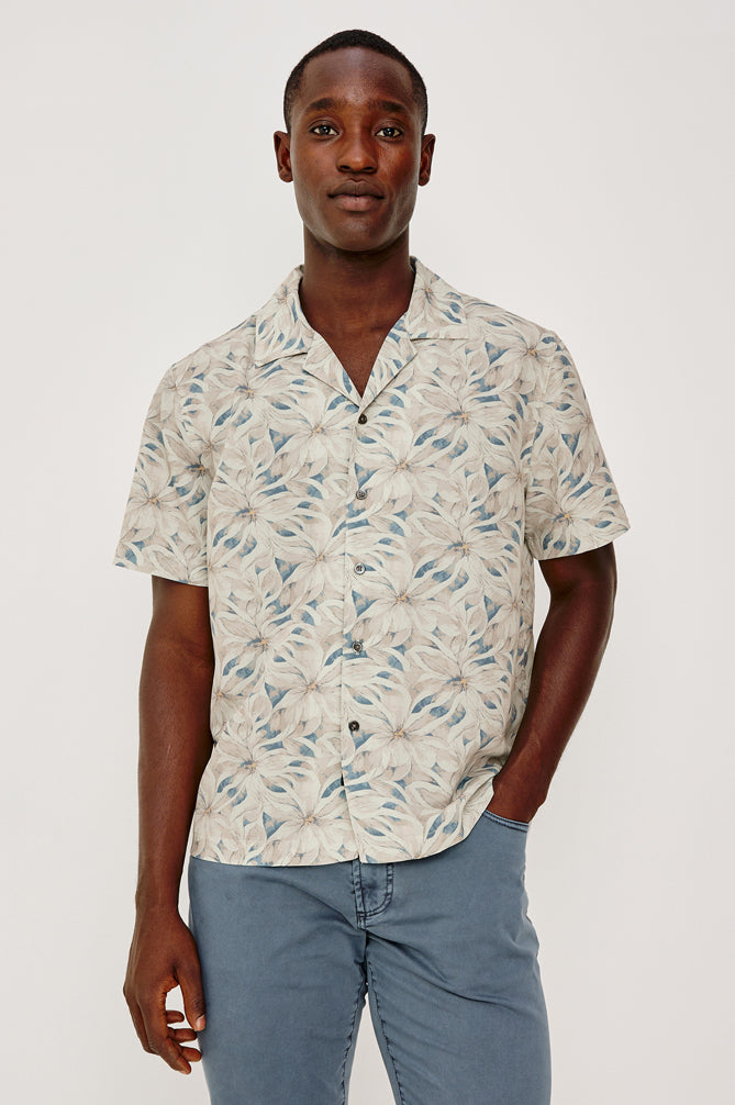 A man stands against a plain background, wearing the Rails Rhodes shirt in Cream Multi with a vintage-inspired leafy pattern and light blue pants. He has one hand in his pocket and maintains a neutral expression.