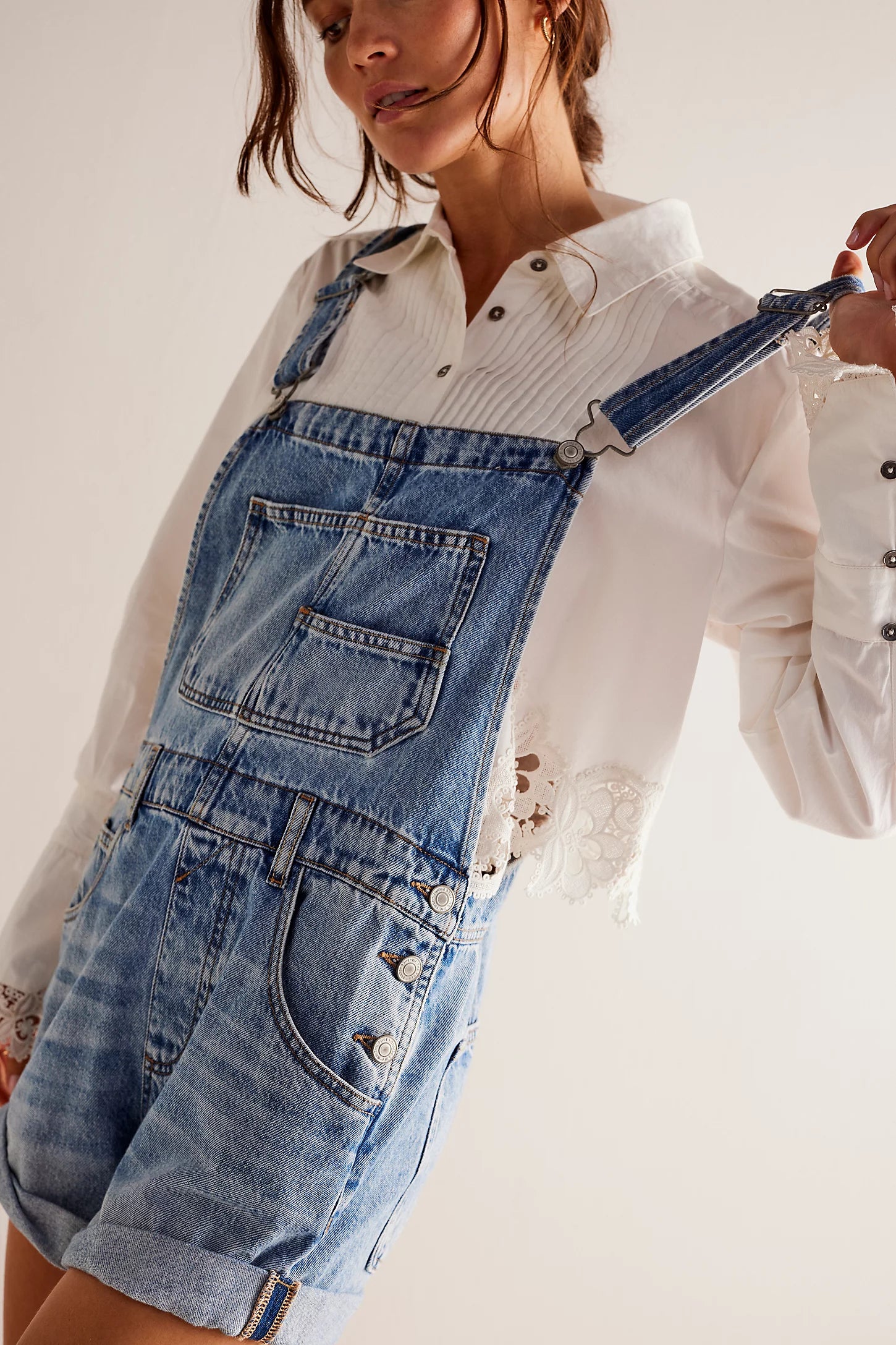 A woman wearing a white long-sleeve lace-trim blouse and Free People Ziggy Shortall blue denim overalls holds one strap in her hand. The image is cropped from her nose down.